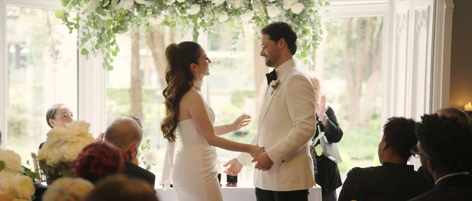 A bride and groom face each other and hold hands, smiling, beneath a canopy of white roses and eucalyptus in front of a large bay window with guests seated either side