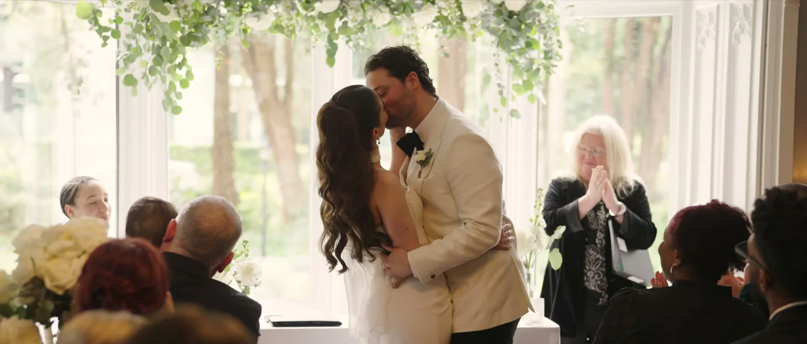 A bride and groom kiss in front of a bay window beneath hanging white flowers as a woman beside them claps and guests look on