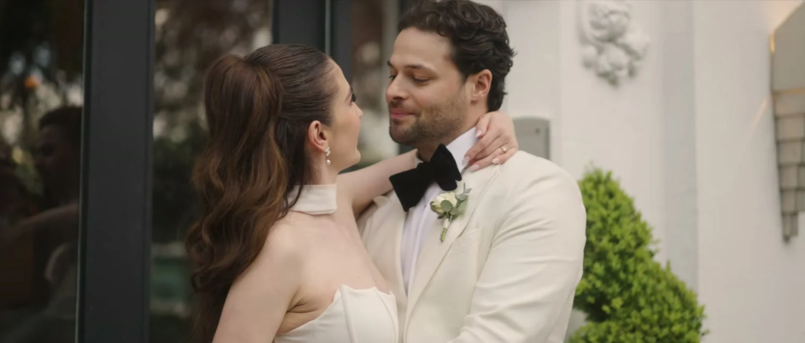 A bride with her arm around her groom's neck smiles up at him beside a white wall with decorative plasterwork and a clipped green shrub