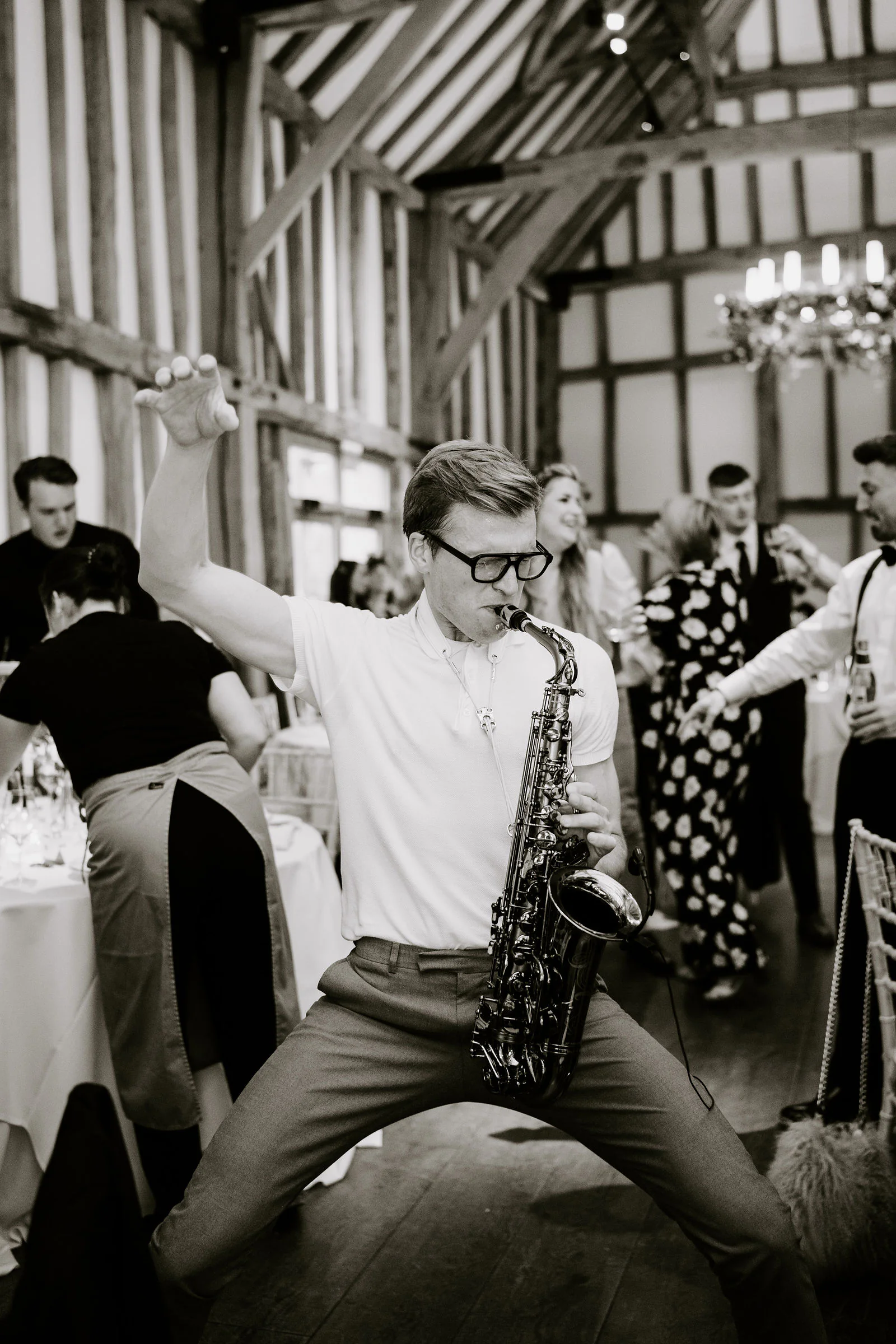 In black and white, a saxophonist in glasses plays with one arm raised and his legs wide among dancing guests inside a timber-framed barn with a chandelier