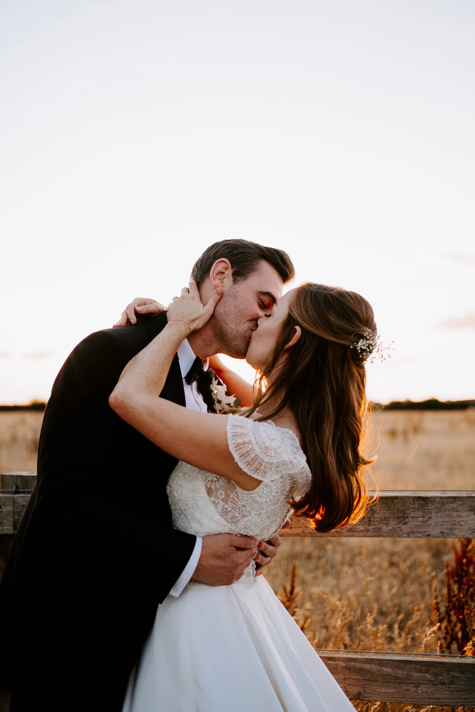 A bride in a lace-sleeved gown and a groom in a dark morning suit kiss as the bride is dipped back, her hand on his neck, against a wooden fence and a golden field in low evening light at Micklefield Hall