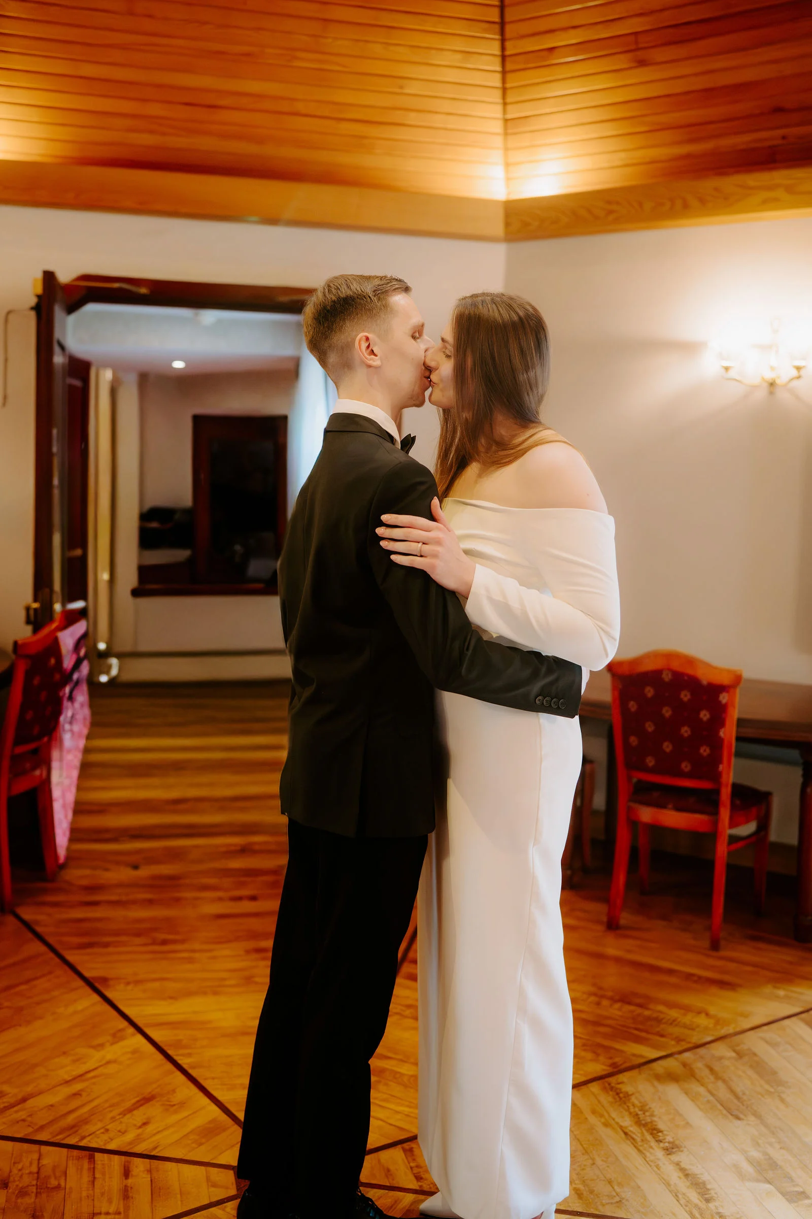 A groom in a black dinner suit and a bride in an off-the-shoulder white gown kiss in a room with a timber ceiling, red upholstered chairs and a polished parquet floor