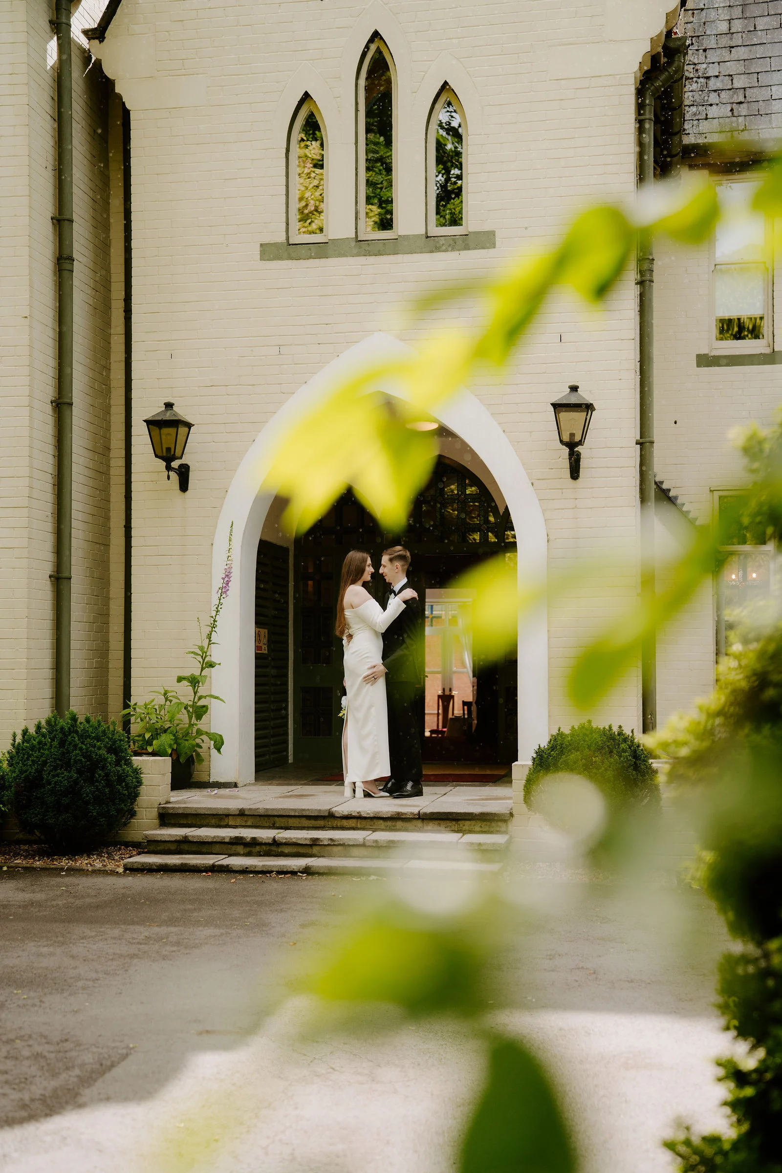A bride in a white gown and a groom in a dark suit stand together in a pointed-arch doorway of a cream-painted house with gothic windows, framed by out-of-focus green leaves