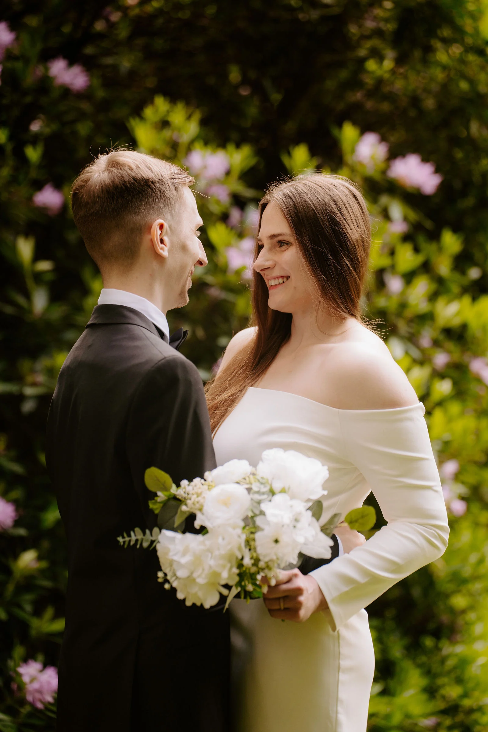 A groom in a black dinner suit and a bride in an off-the-shoulder white gown holding a bouquet of white flowers smile at each other among pink rhododendron blooms in a garden