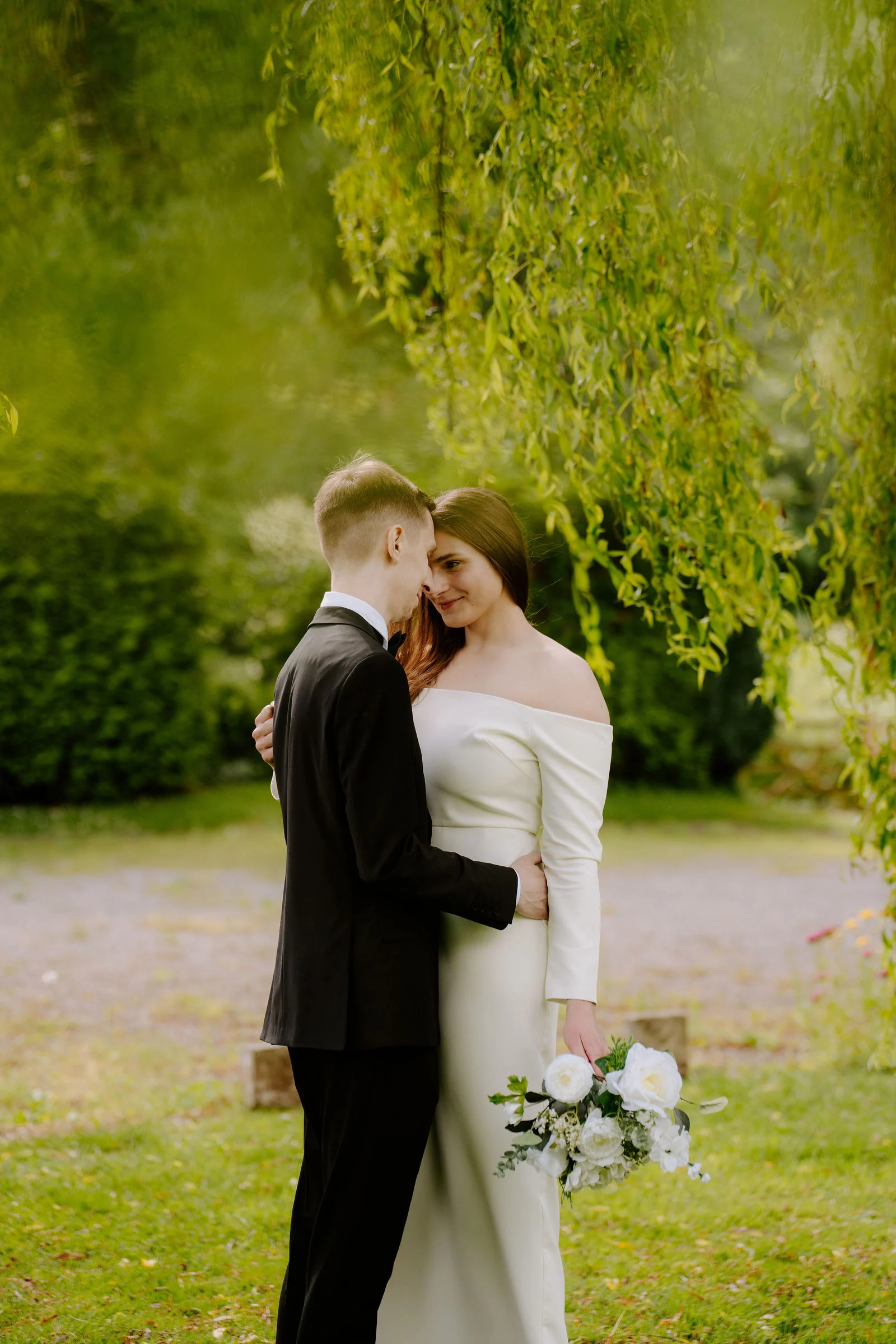A groom in a dark suit holds his bride in a white gown close on a lawn beneath the trailing branches of a weeping willow, her bouquet of white flowers in her hand