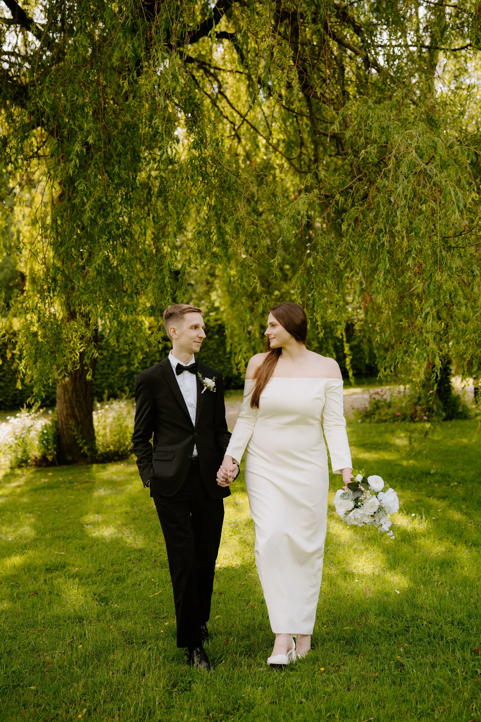 A groom in a black tuxedo and bow tie and a bride in an off-the-shoulder cream gown holding a bouquet of white flowers walk hand in hand on a lawn beneath a weeping willow