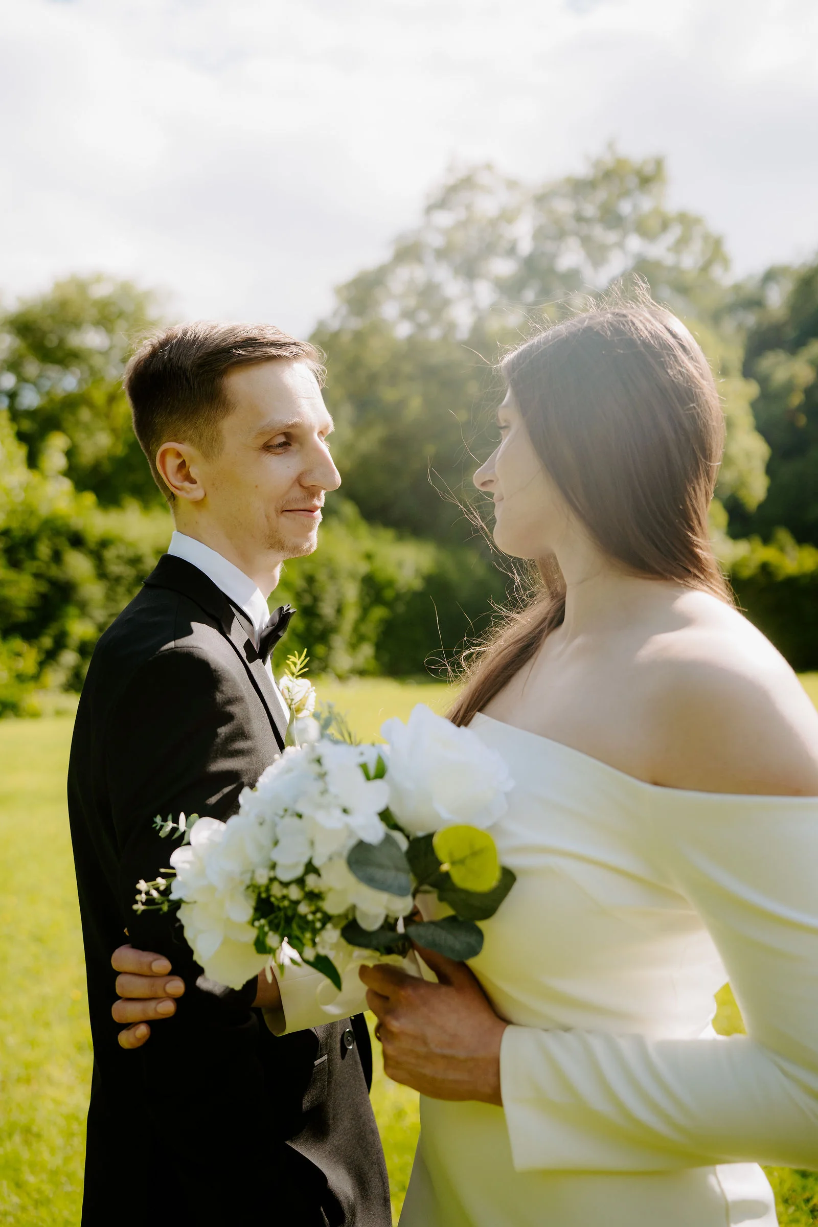 A groom in a black tuxedo and a bride in an off-the-shoulder cream gown holding a bouquet of white flowers turn to face each other in bright sun, with green trees and a hedge behind them