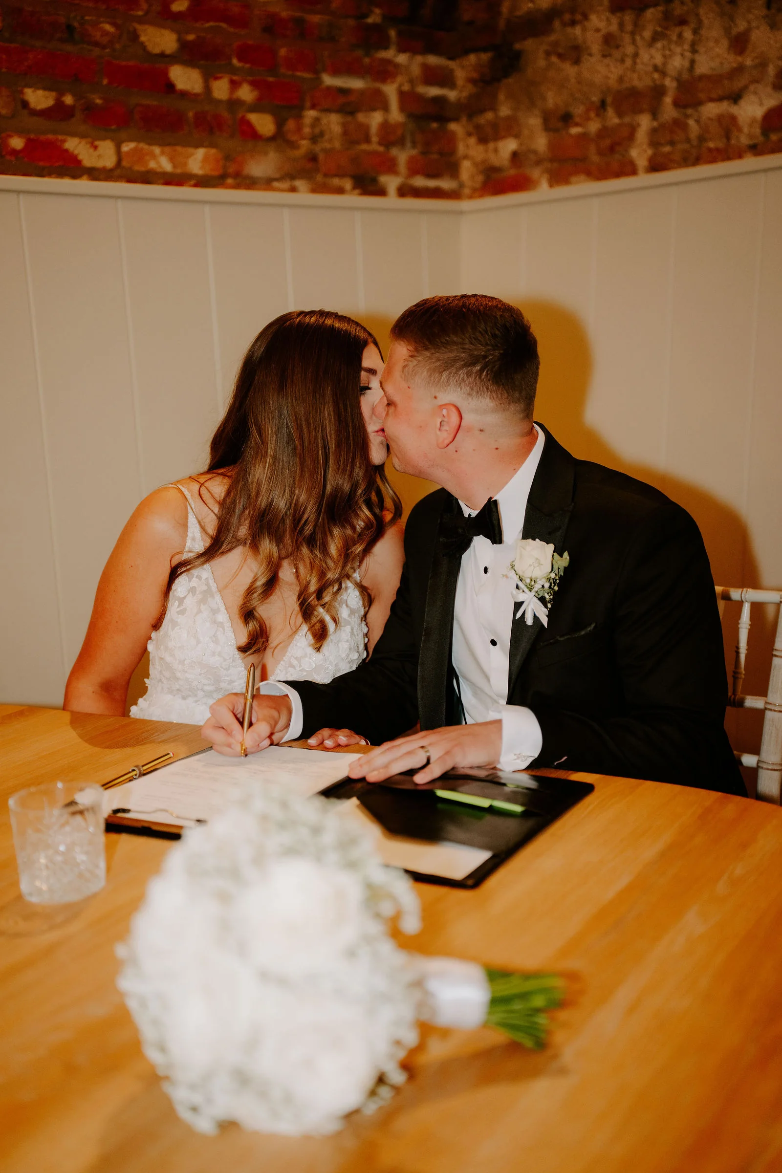 A groom in black tie kisses his bride at a wooden table where they are signing the register, with a bouquet of white roses in the foreground and an old brick wall behind them