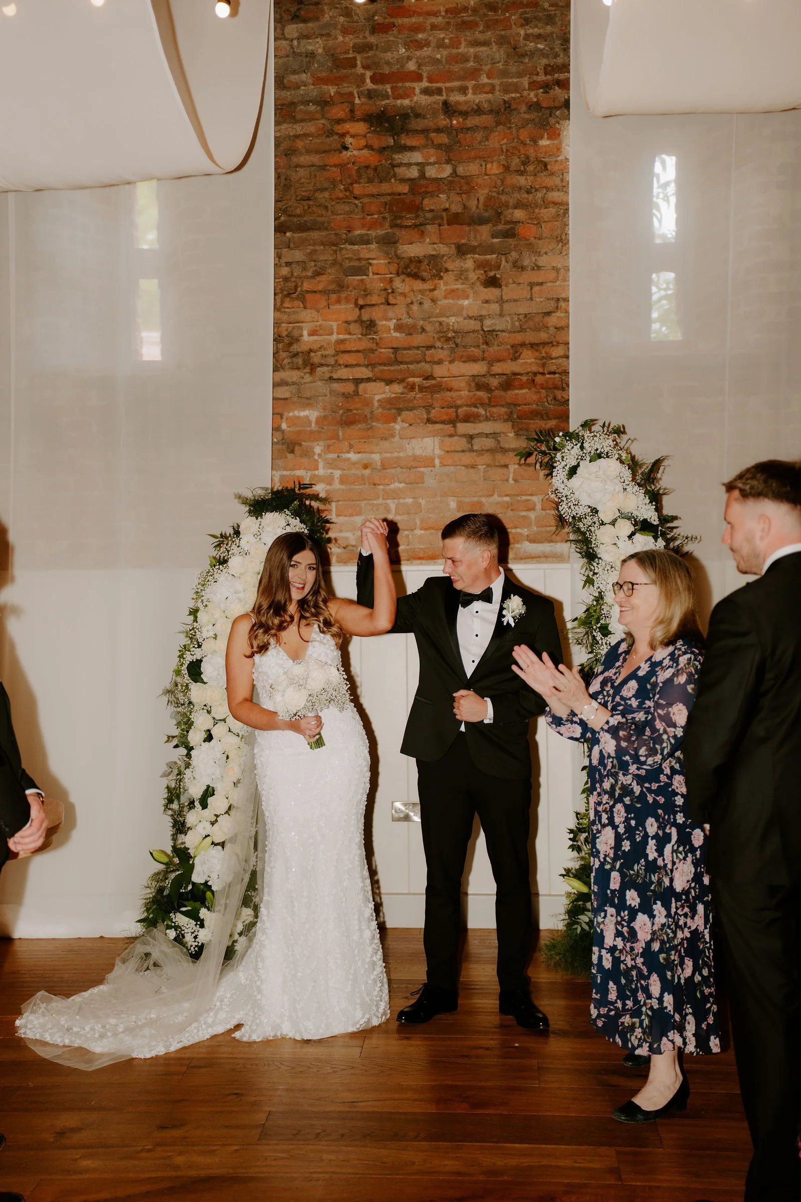 A bride in a lace gown with a long veil and a groom in black tie stand between two white floral arches in front of a brick chimney breast and sheer drapes in a timber-floored barn, the groom raising the bride's hand while guests clap