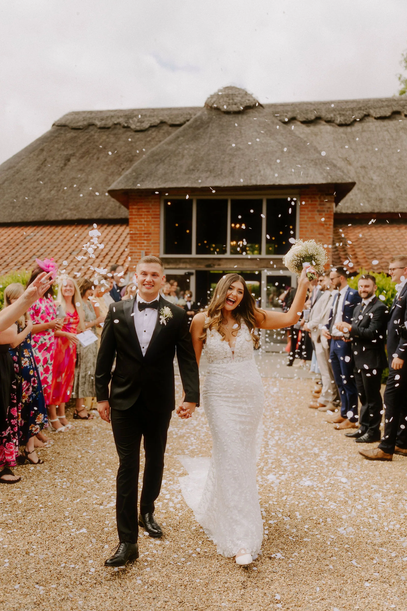A groom in black tie and a bride in a lace gown holding a bouquet walk hand in hand along a gravel path between lines of guests throwing confetti, in front of a thatched barn with a tall glazed gable