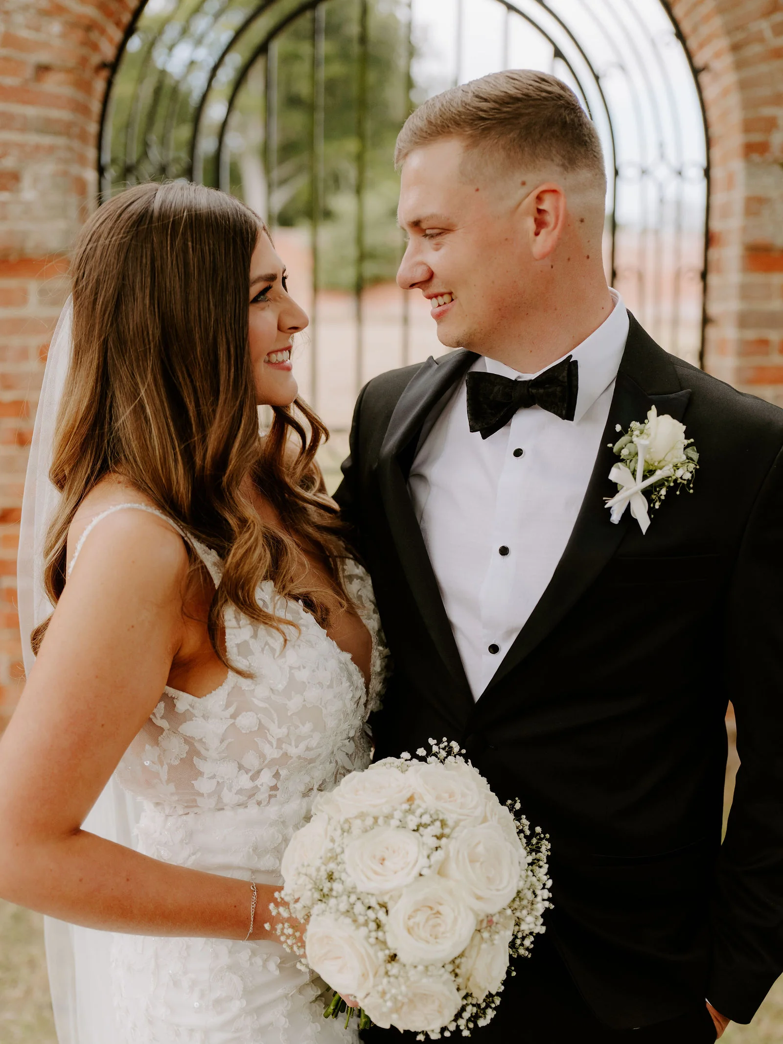 A close portrait of a smiling bride with a veil holding a bouquet of white roses and a groom in a bow tie, framed by an arched iron window in an old brick wall