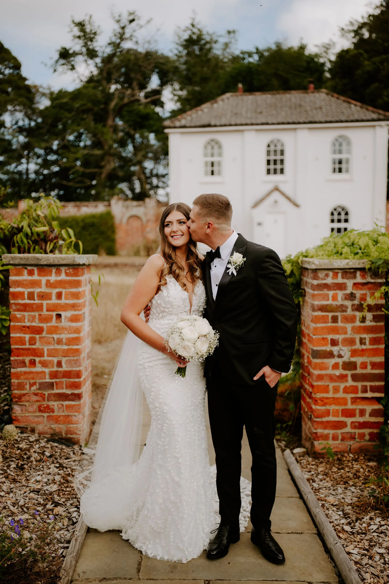 A groom kisses the cheek of his smiling bride, who holds a bouquet of white roses, between two brick gate piers in a walled garden, with a white two-storey building with arched windows behind them