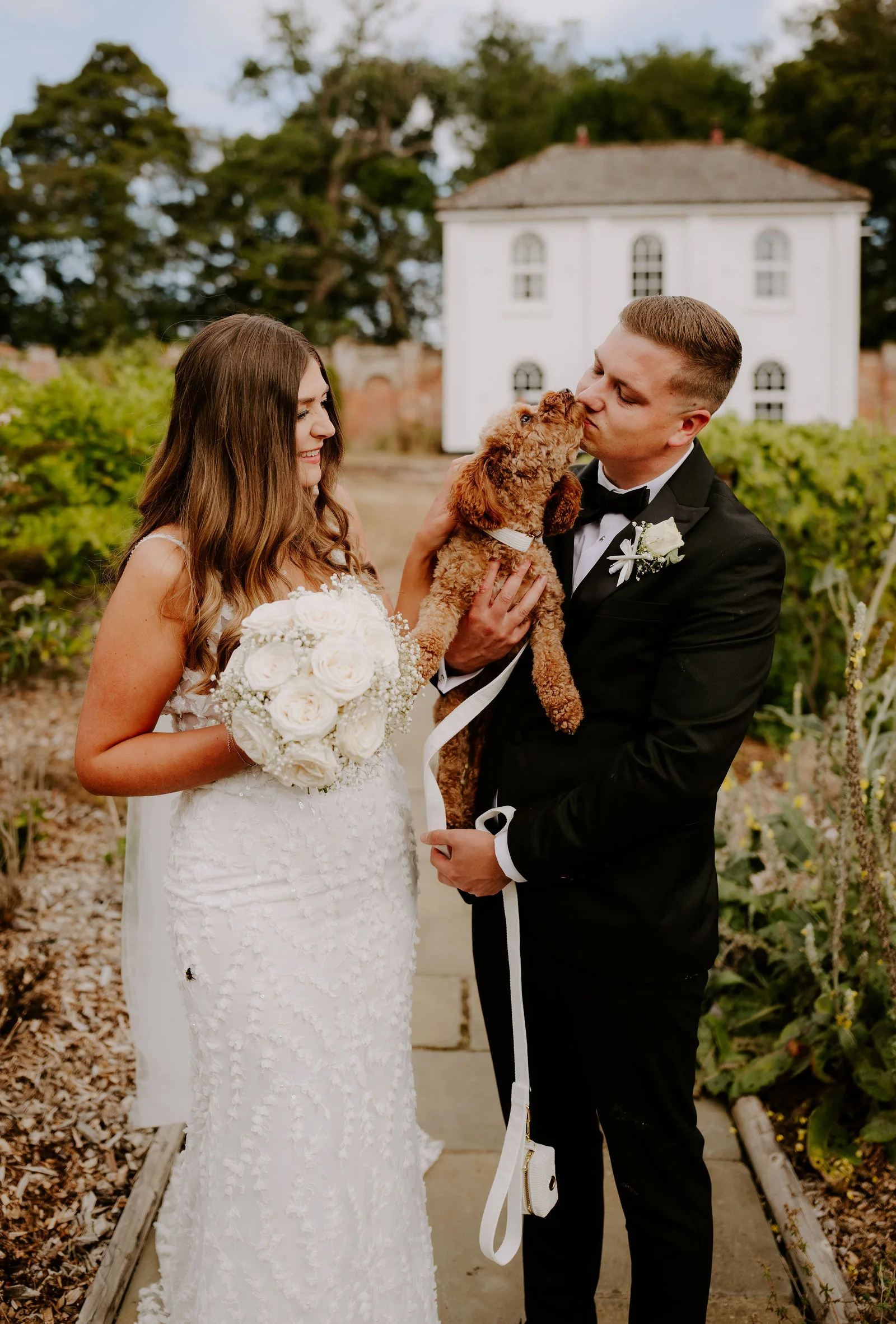 A bride holding a bouquet of white roses and a groom in black tie hold a small curly-haired brown dog on a lead between them, and the groom kisses the dog, in a walled garden with a white building behind