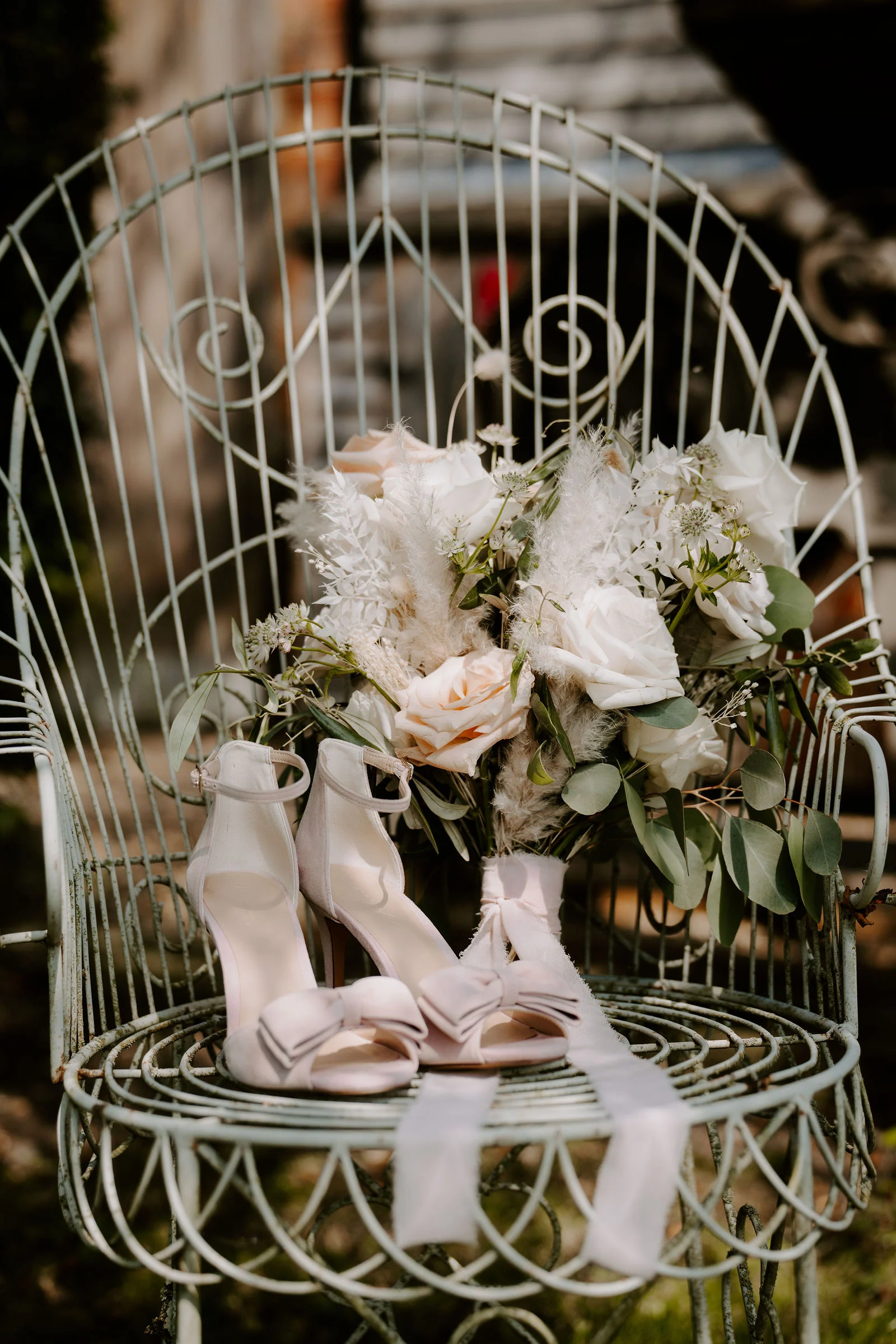 A pair of blush heeled sandals with bows and a bouquet of cream roses and dried grasses rest on a pale green wire chair outdoors, with soft greenery behind.