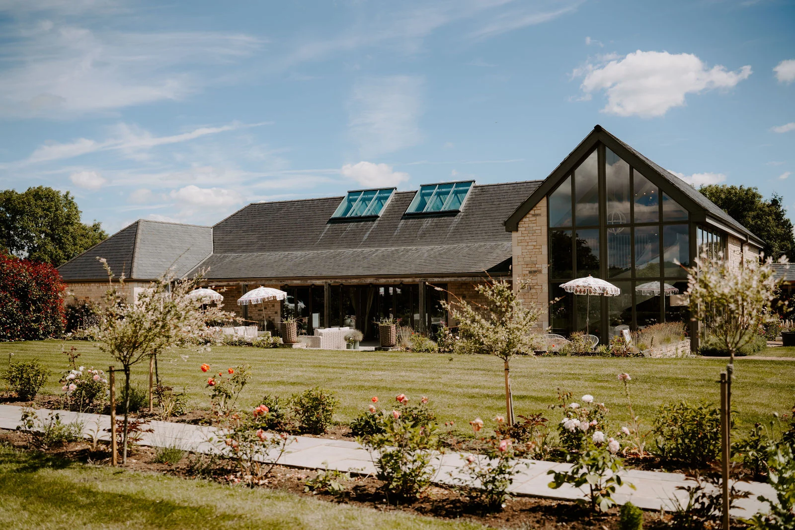 A long, low barn with a slate roof, skylights and a tall glass gable end stands beyond a sunlit lawn edged with rose bushes, with white parasols on the terrace and a blue sky with light cloud above.