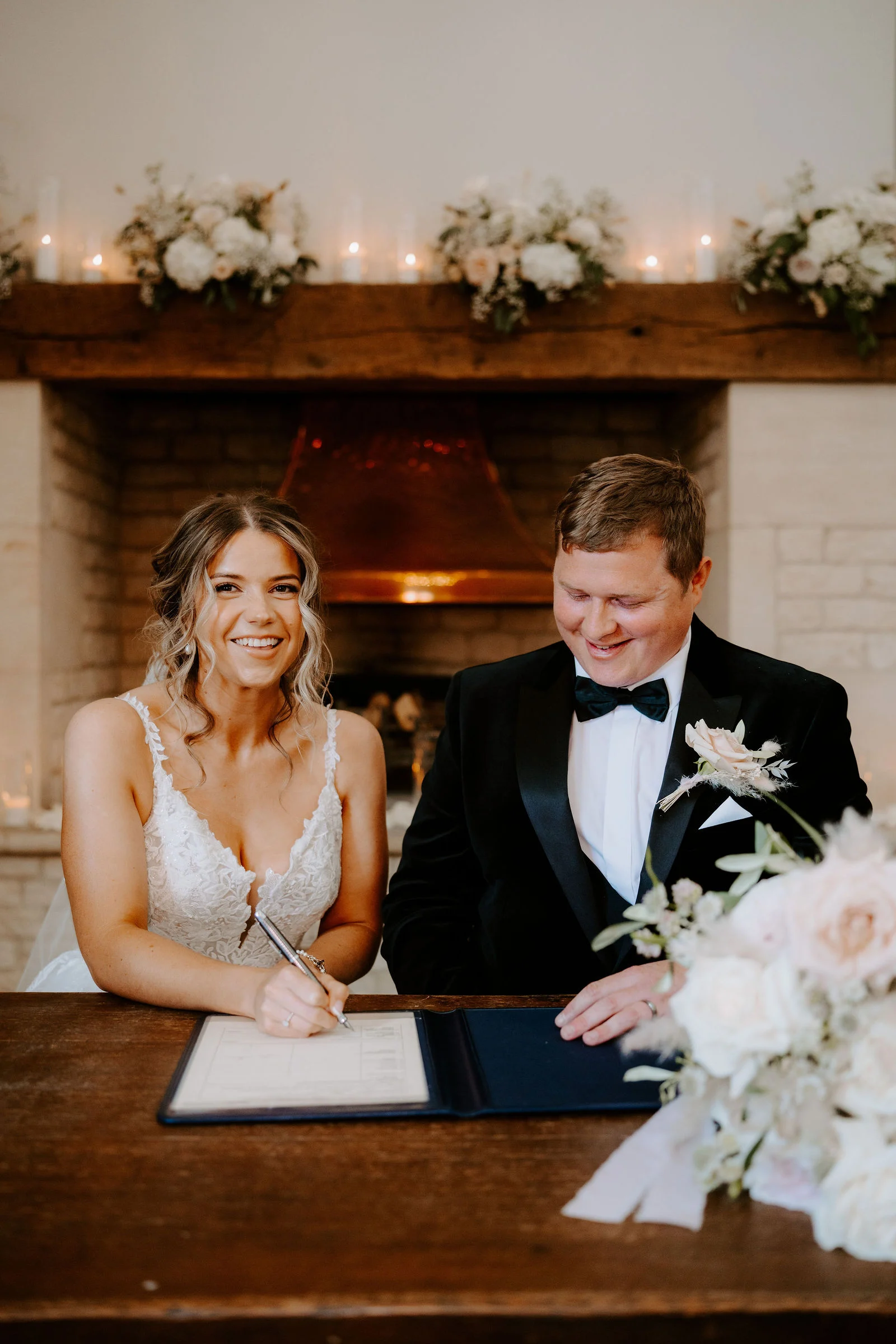A smiling bride in a lace-strap gown signs the register at a wooden table while the groom in a black dinner jacket looks down with a smile, with a stone fireplace topped with flowers and candles behind them.