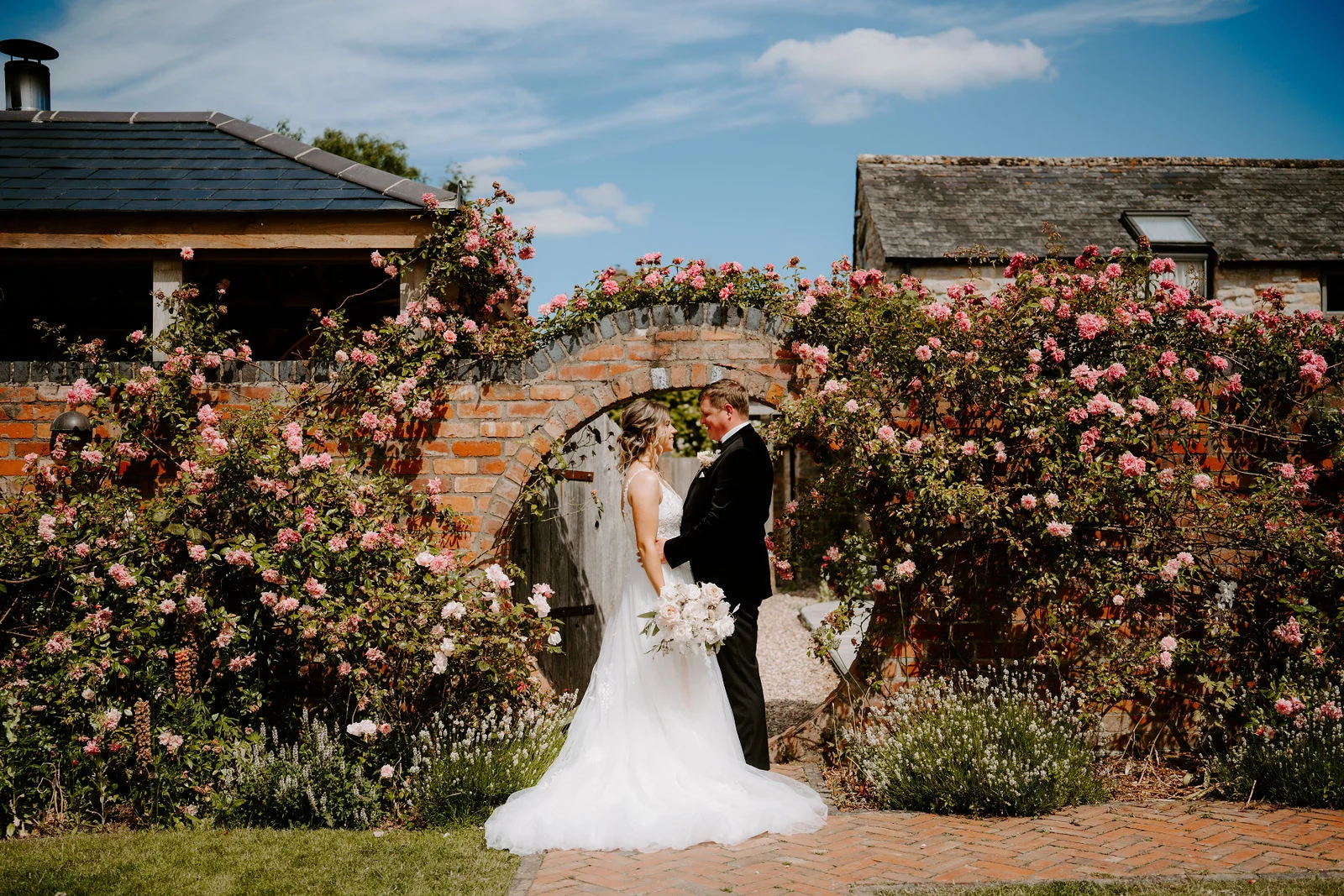 A bride in a long white gown and a groom in a black dinner jacket stand face to face in a round brick archway covered in pink roses, with slate-roofed barns behind them and a bright blue sky above.