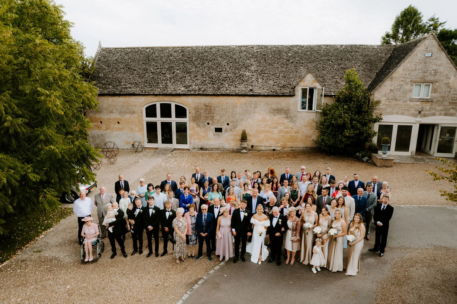From above, a large wedding party gathers on a gravel courtyard in front of a long stone barn with a tiled roof and tall arched window