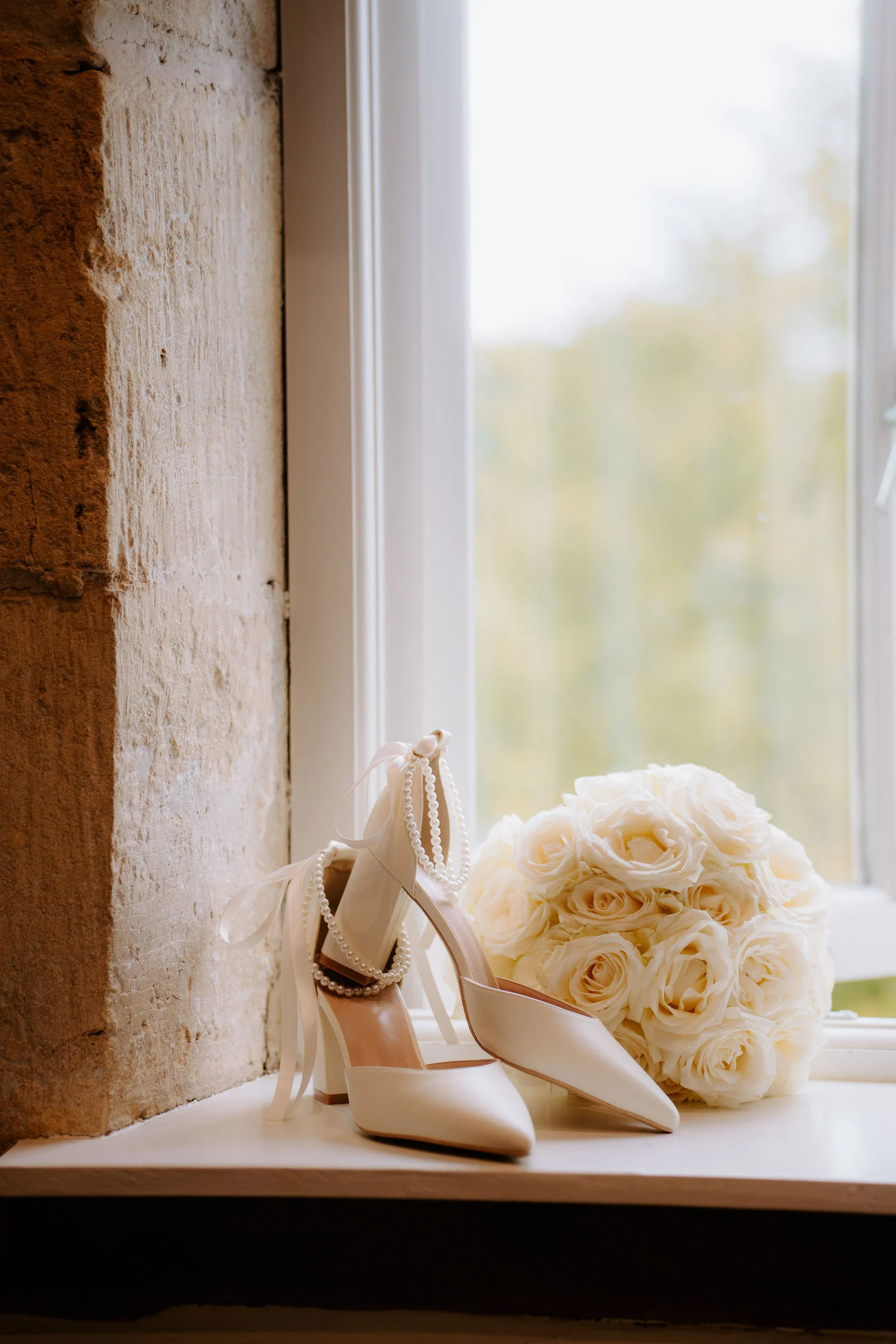 A pair of ivory block-heeled shoes with pearl ankle straps sits on a deep stone window sill next to a round bouquet of cream roses, with soft green trees blurred through the glass.
