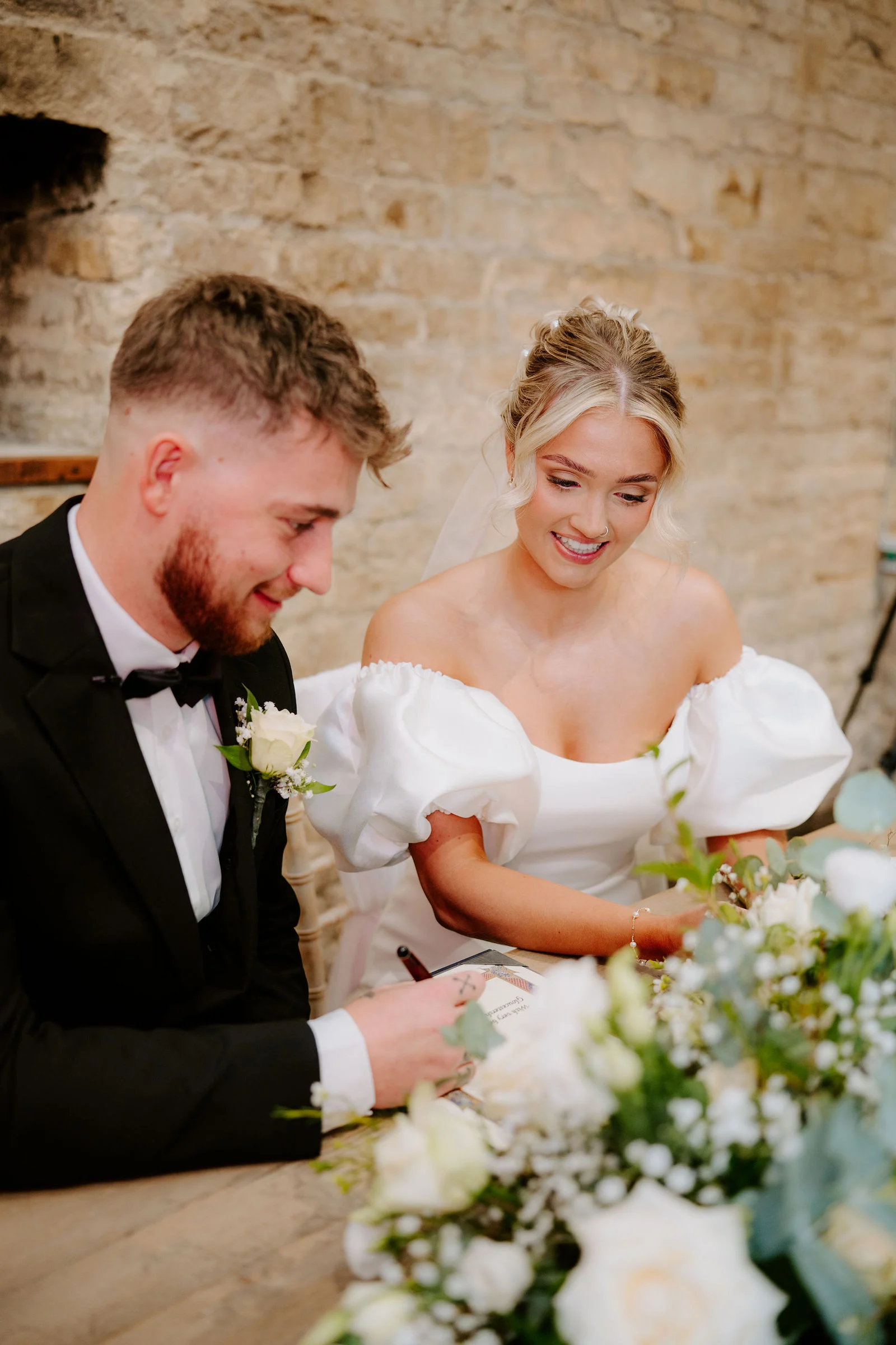 A bearded groom in a black suit and a bride in a white dress with puffed sleeves smile as they look down to sign the register, with a bunch of white and green flowers in front of them and a stone wall behind.