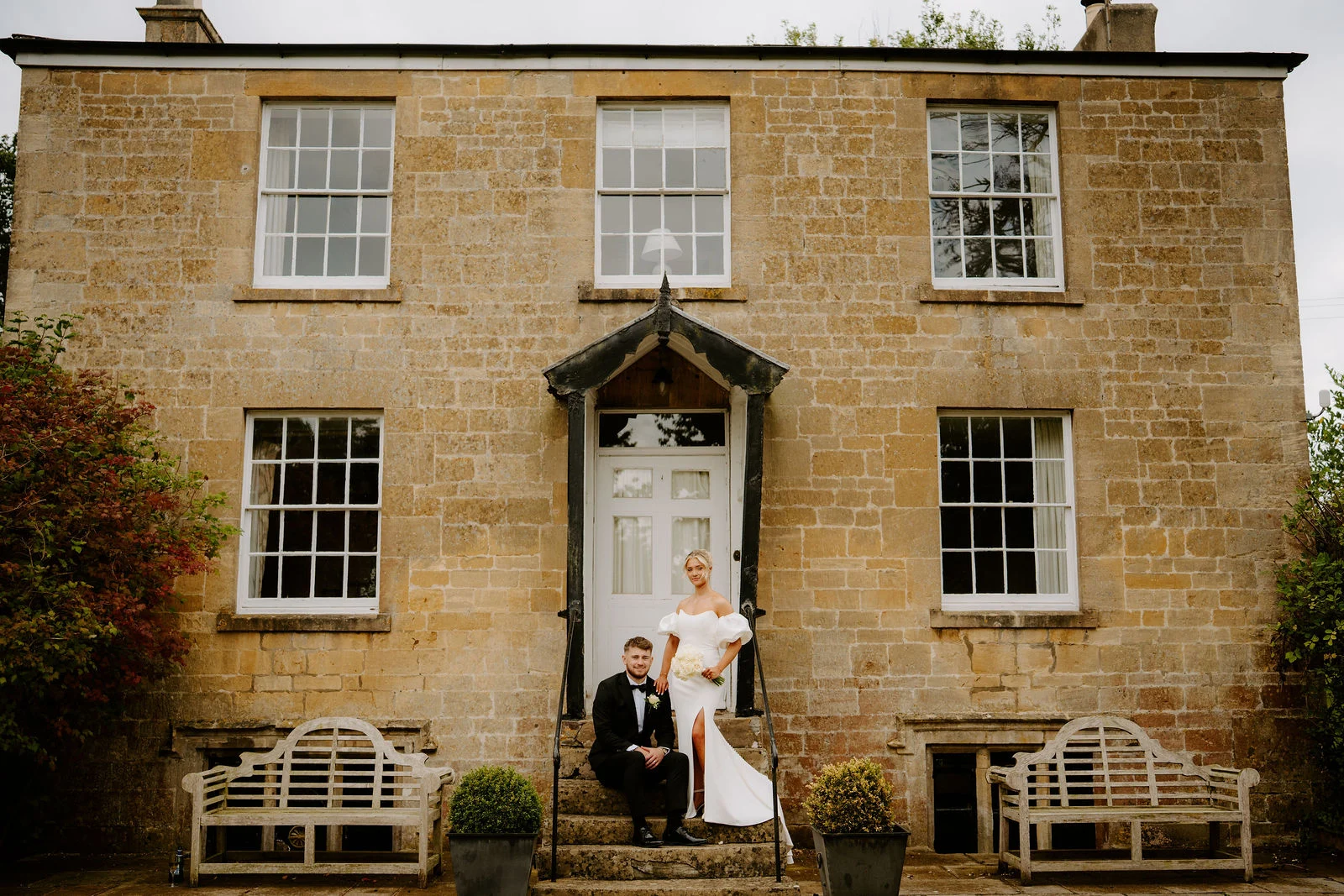 A groom in a black dinner suit sits on the stone steps of a honey-coloured farmhouse while the bride in a white off-the-shoulder dress stands beside him at the white front door, with garden benches either side and a lawn in front.