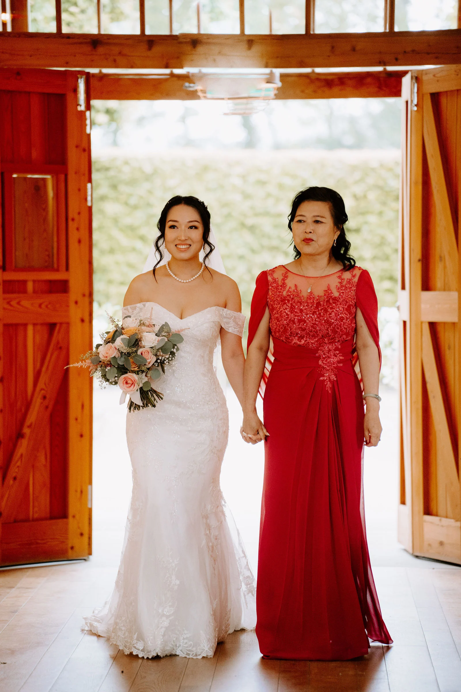 A bride in a fitted lace gown holding a soft pink bouquet walks hand in hand with a woman in a red dress through open wooden barn doors, the bride smiling.