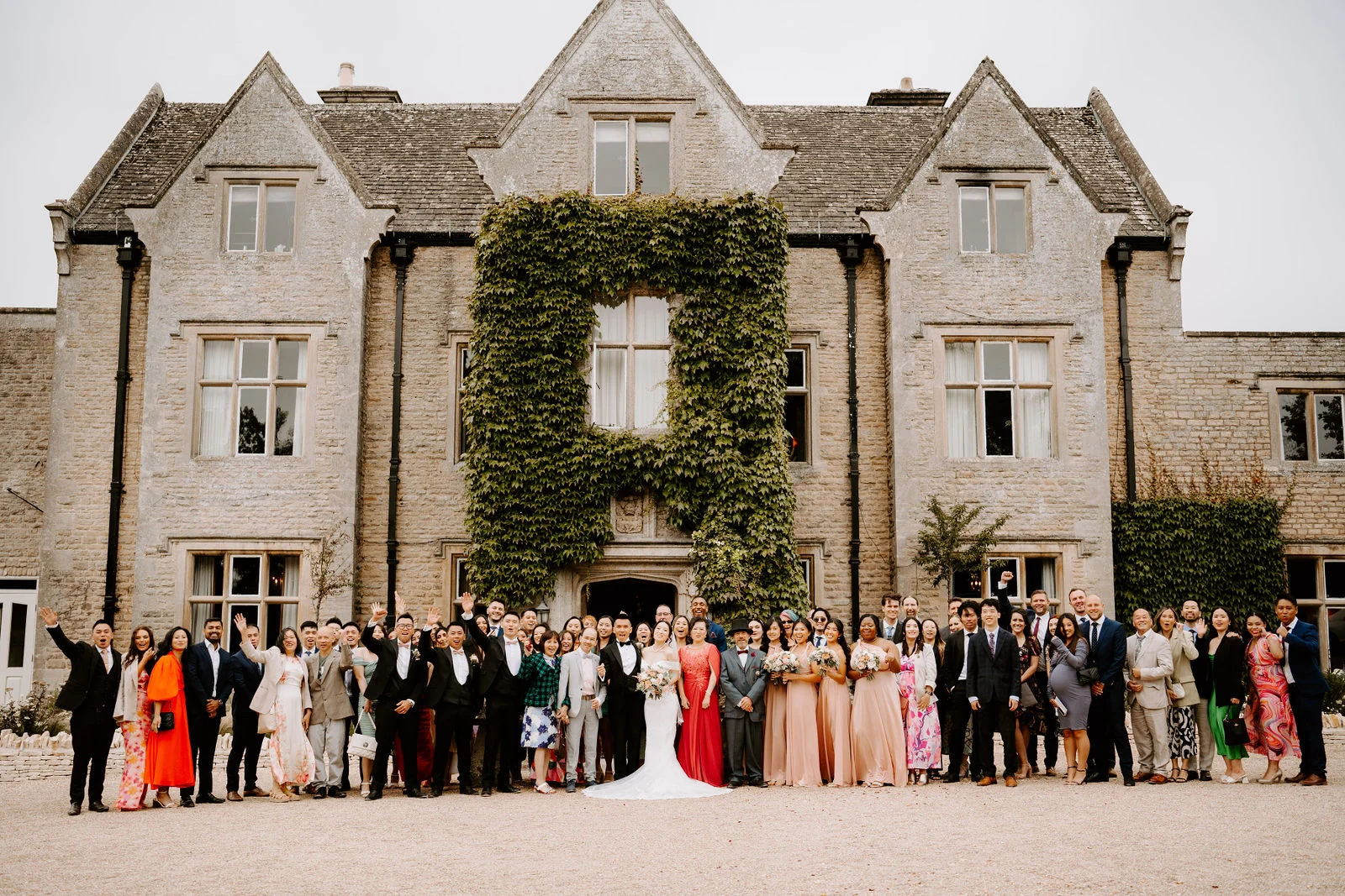 A large wedding party stands in a long row on the gravel in front of the ivy-covered stone manor, the bride in white at the centre and several guests waving and cheering.