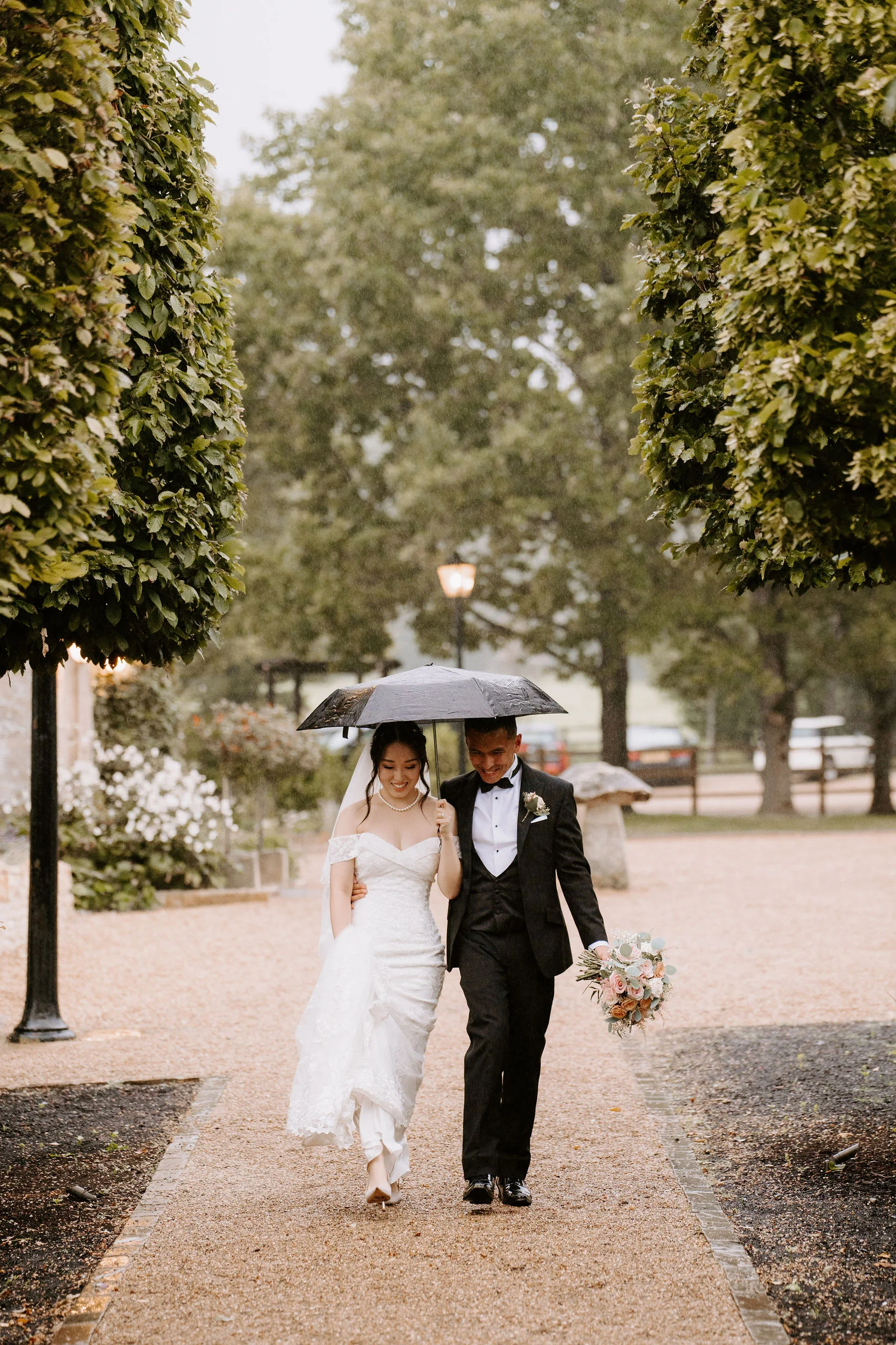 A bride and groom walk arm in arm along a gravel path between clipped trees, sharing a black umbrella in the rain and smiling as they look down.