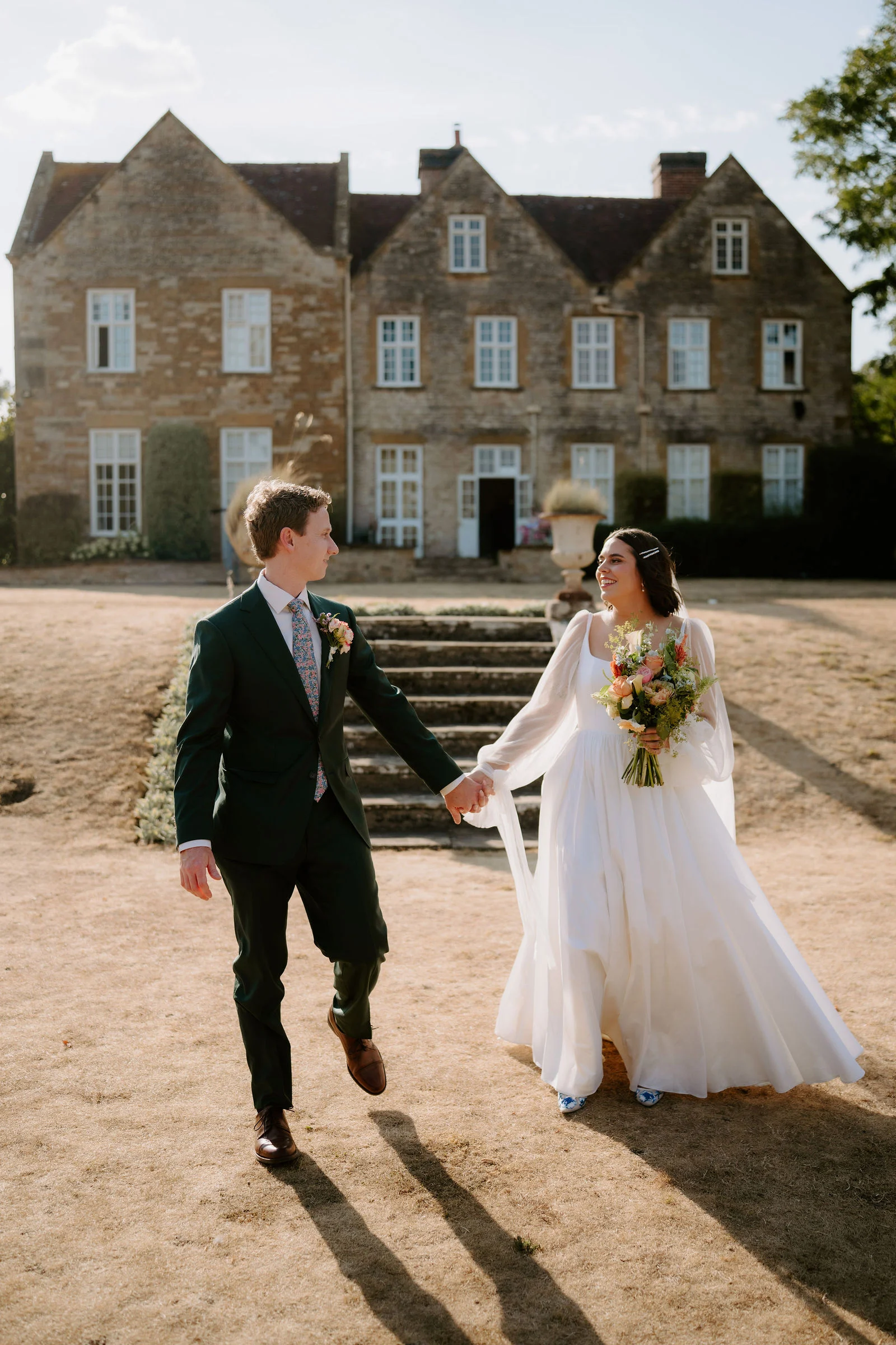 A groom in a dark green suit and a bride in a long white dress with a bouquet walk hand in hand across a dry lawn, smiling at each other, with a stone manor house and steps behind them.