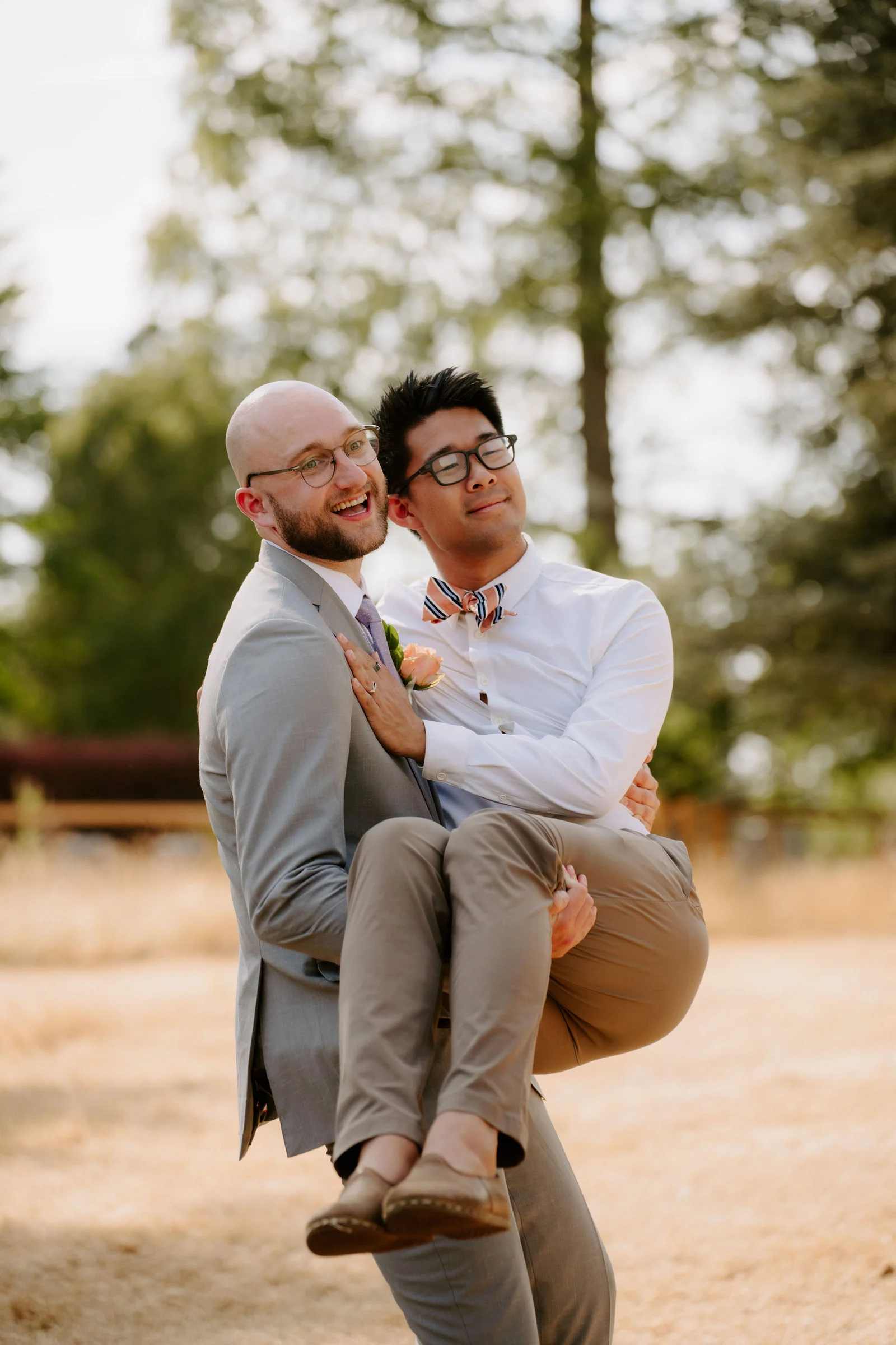 Two men laugh together outdoors as one in a grey suit lifts the other, in a white shirt and bow tie, into his arms, with trees behind them.