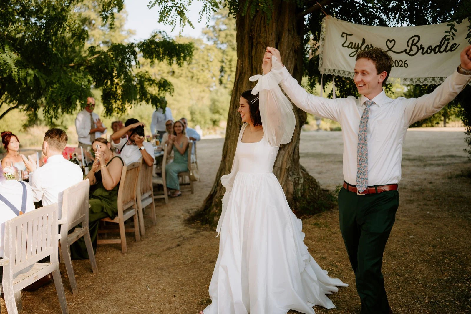 A bride in a white dress with sheer sleeves and a groom in a white shirt and tie raise their arms as they walk past a long outdoor dining table where guests sit under trees.