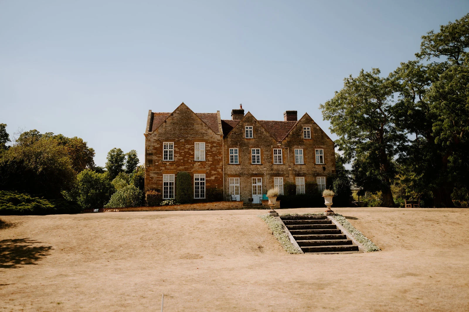 A honey-coloured stone manor house with gabled roofs stands at the top of a stone staircase across a dry, sunlit lawn under a clear blue sky.