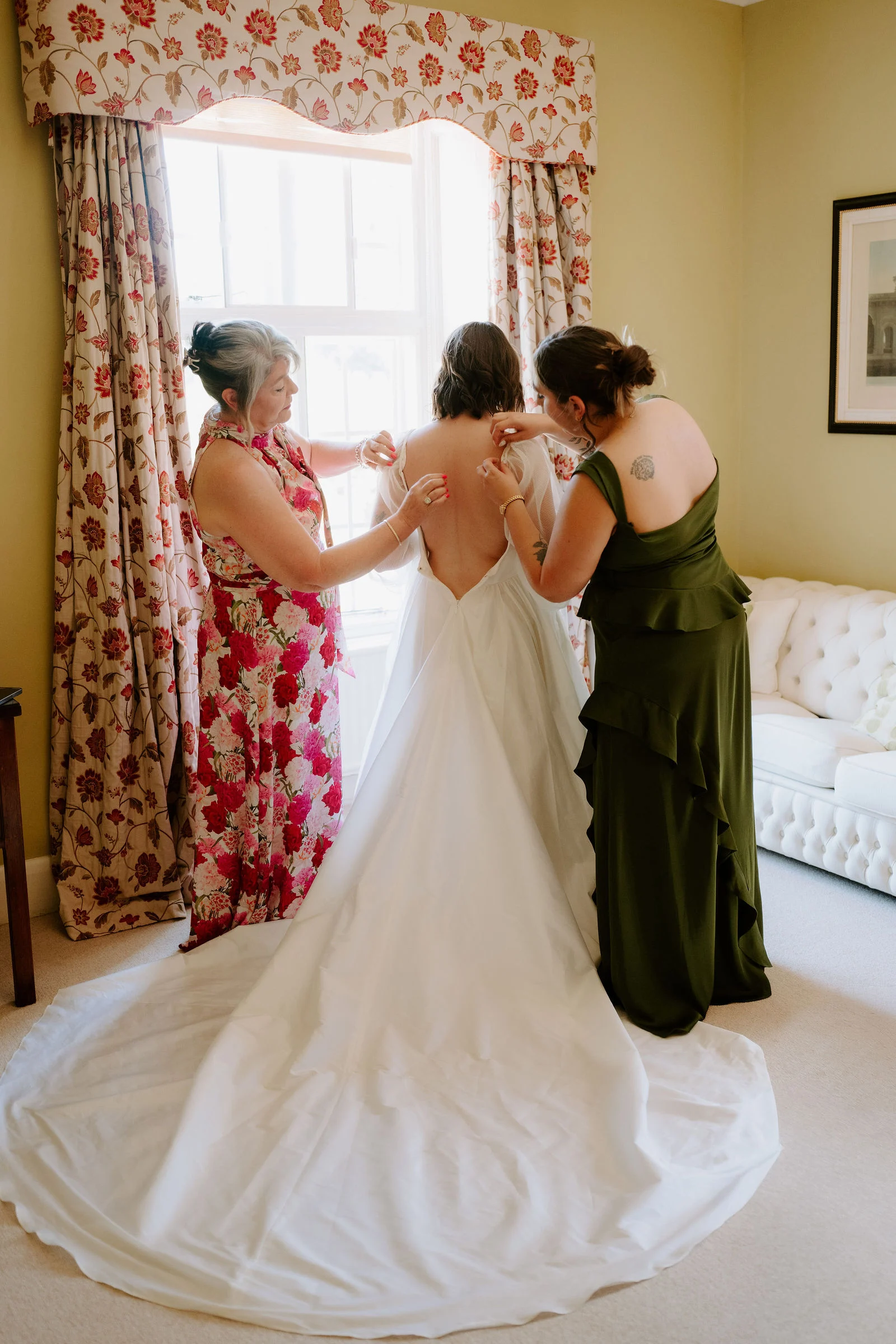Two women in a floral dress and an olive green dress fasten the back of a wedding dress with a long train beside a tall window with floral curtains.