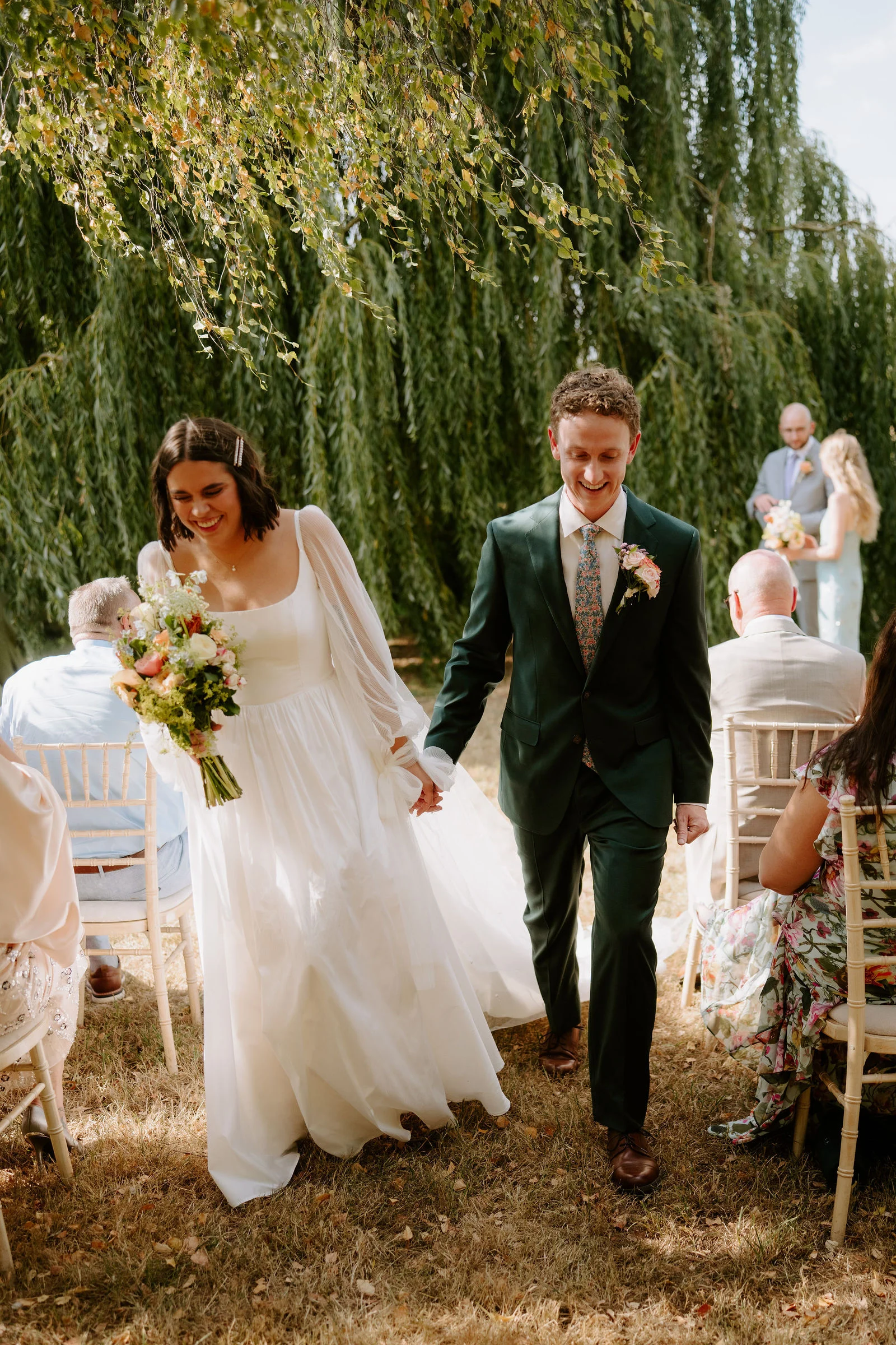 A bride in a white dress holding a bouquet and a groom in a dark green suit walk hand in hand along a grass aisle, both smiling, past seated guests beneath a weeping willow.