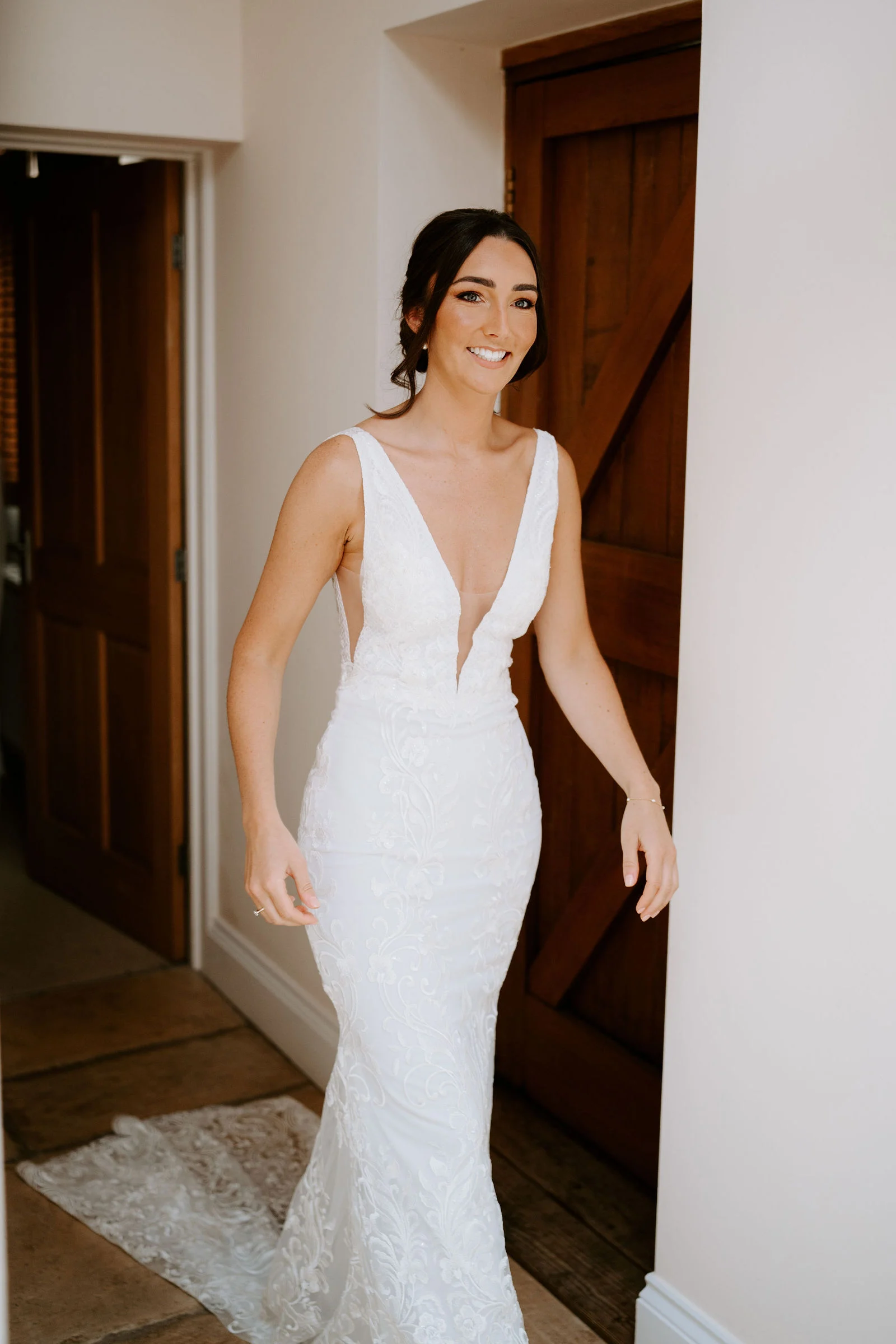 A smiling bride in a fitted white gown with a plunging neckline steps through a wooden door into a white-walled room