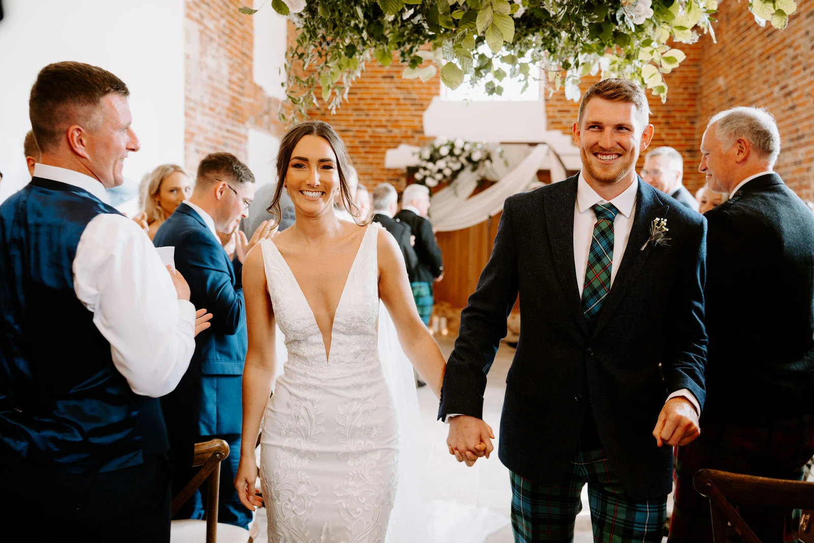 A smiling bride in a white dress and a groom in a navy jacket and tartan trousers walk hand in hand down the aisle of a brick barn between seated guests.