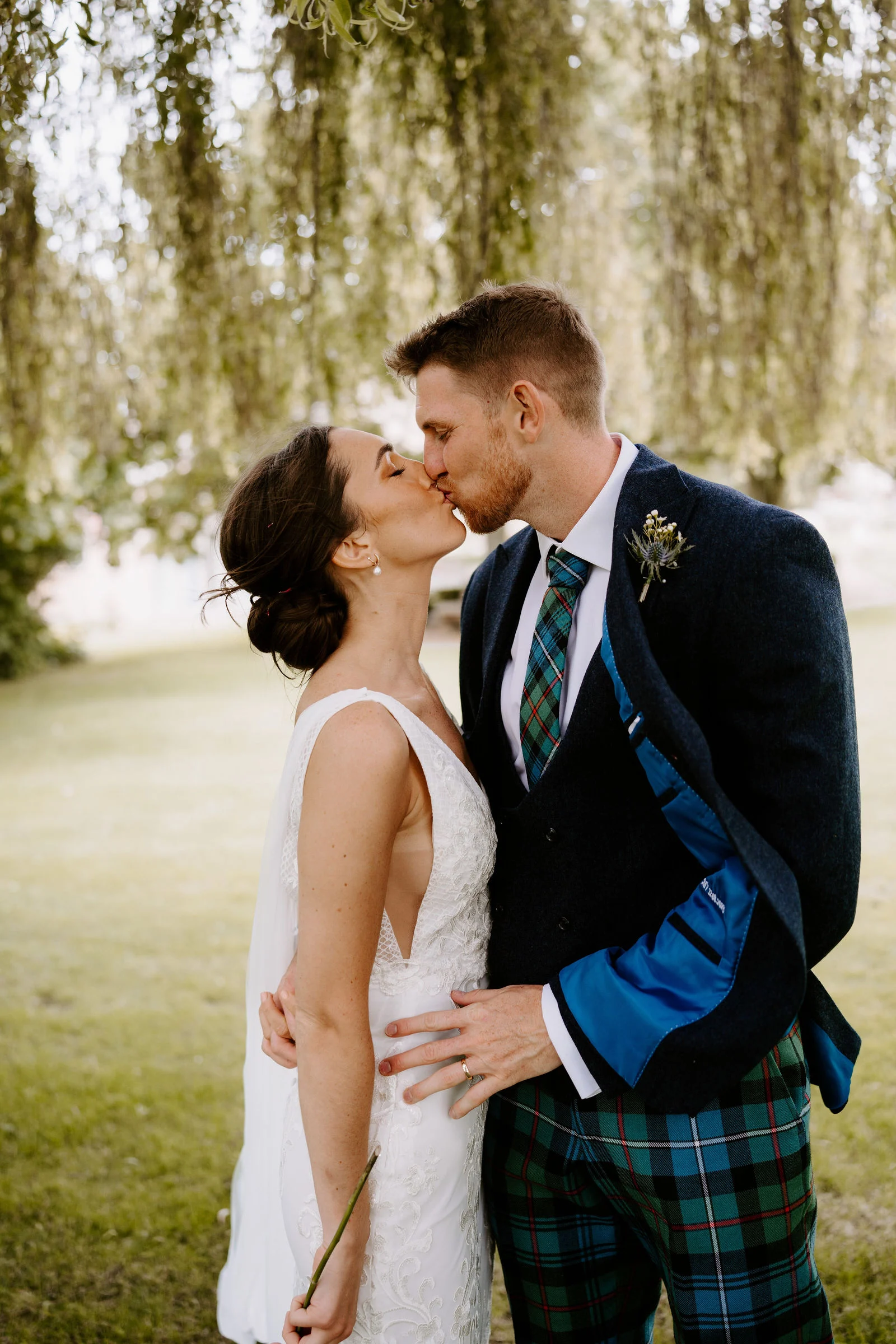A bride in a white dress and a groom in a navy jacket and tartan trousers kiss on a lawn beneath the hanging branches of a weeping willow.