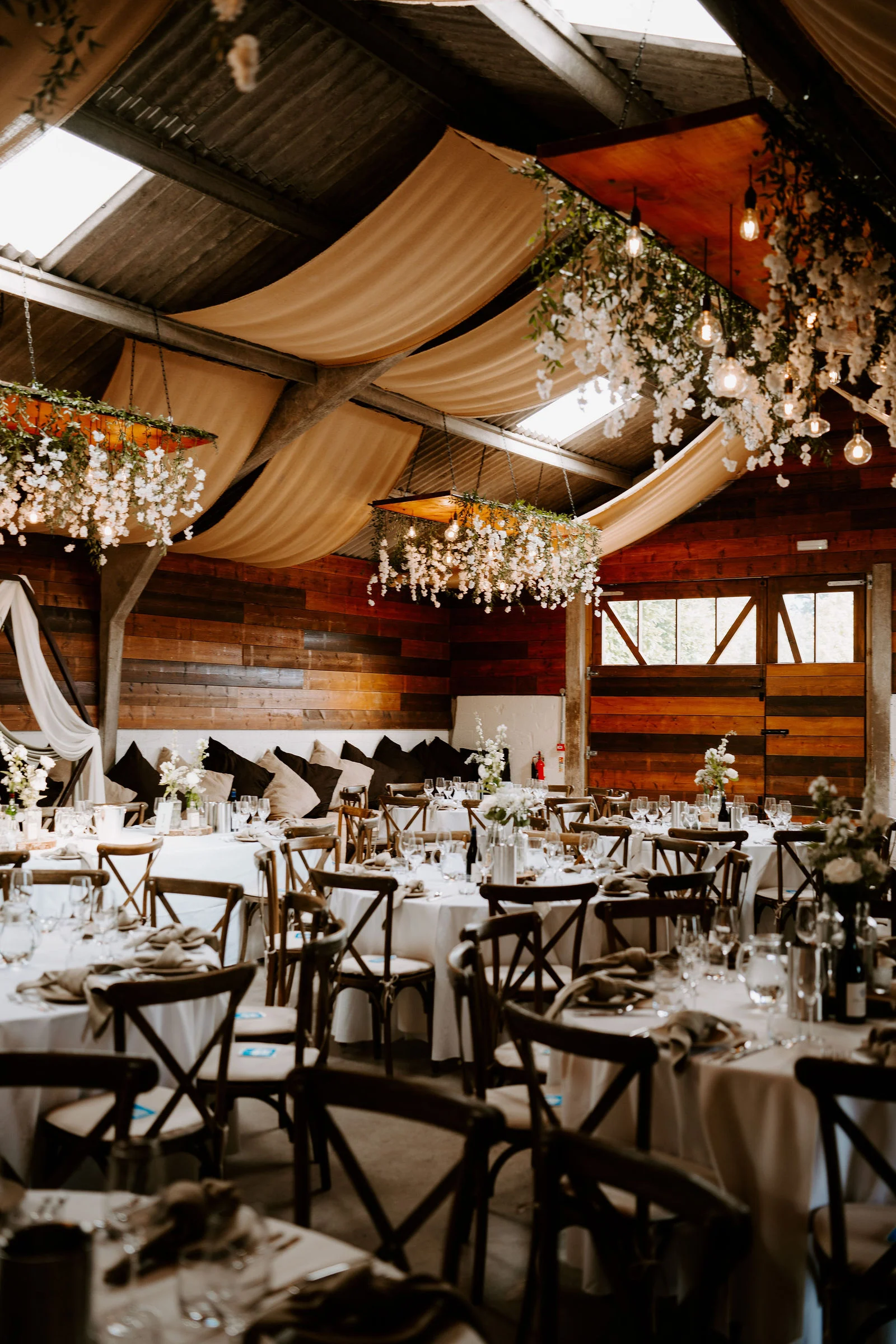 A barn laid out for a wedding meal, with round tables, wooden cross-back chairs, draped cream fabric across the ceiling and hanging white flowers.