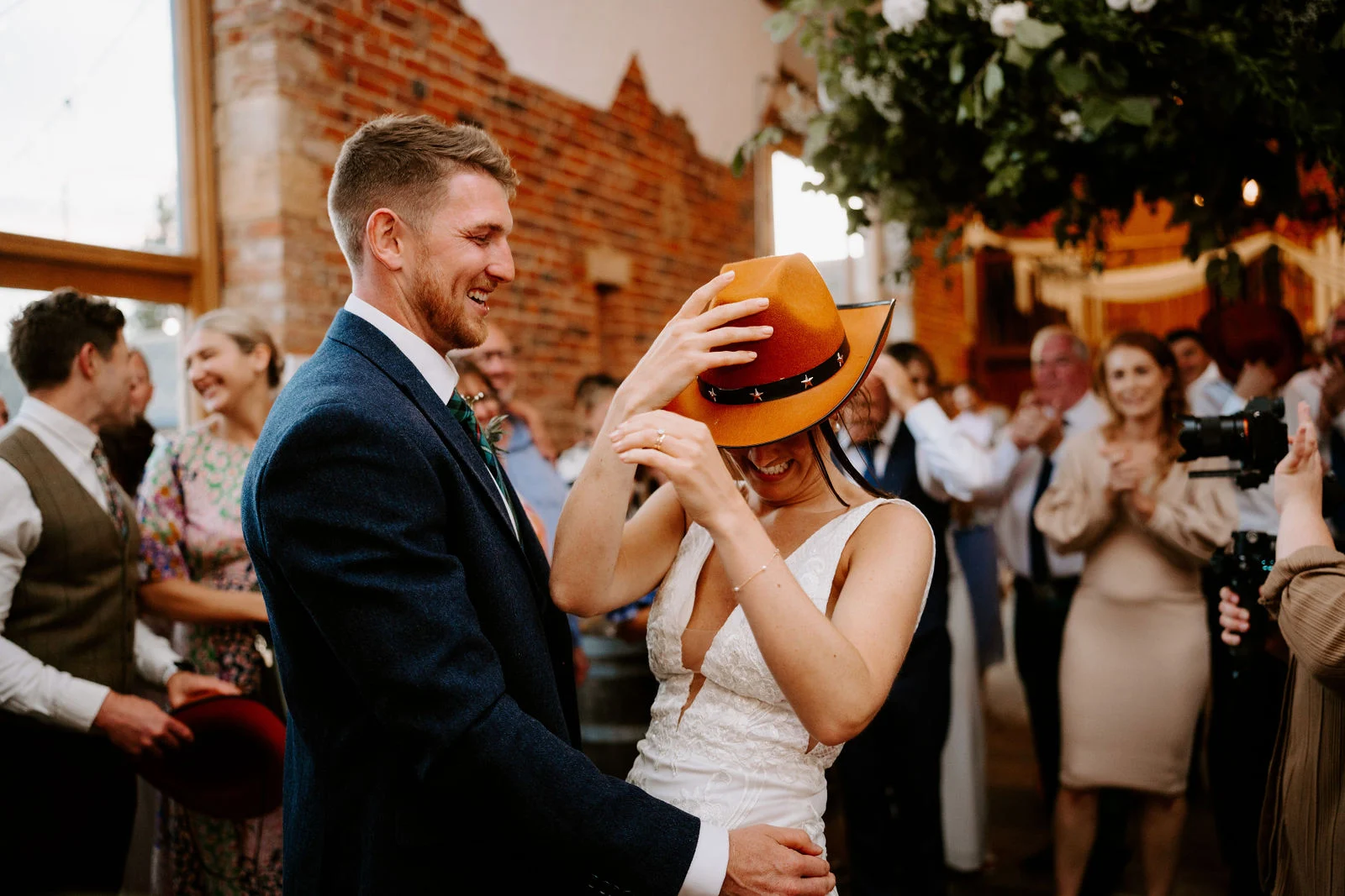 A groom in a navy suit laughs as a bride in a white dress pulls an orange cowboy hat down over her face while they dance, with guests clapping behind them in a brick barn.