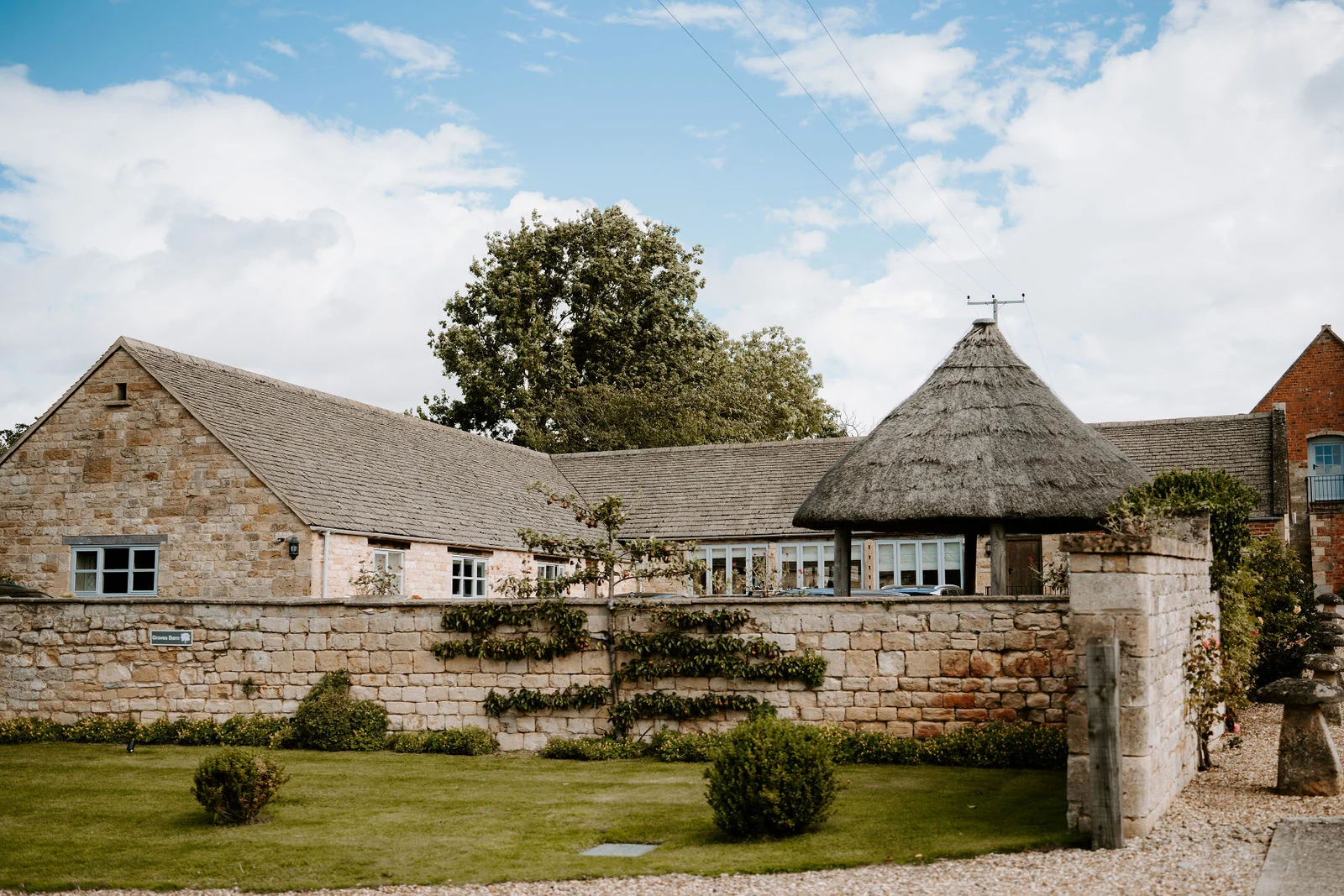 A low stone building with a thatched round roof sits behind a clipped lawn and a stone wall, under a bright blue sky with white clouds.