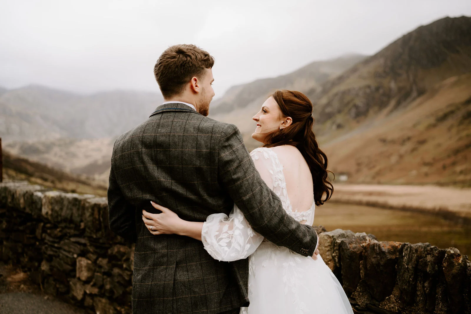 A groom in a grey check jacket holds his bride at the waist as she looks over her shoulder at him, with a stone wall and mountains behind them near Hafod Farm