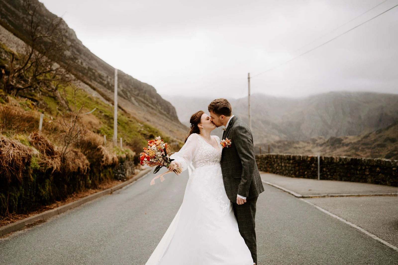 A bride and groom kiss on an empty mountain road with steep slopes rising either side near Hafod Farm