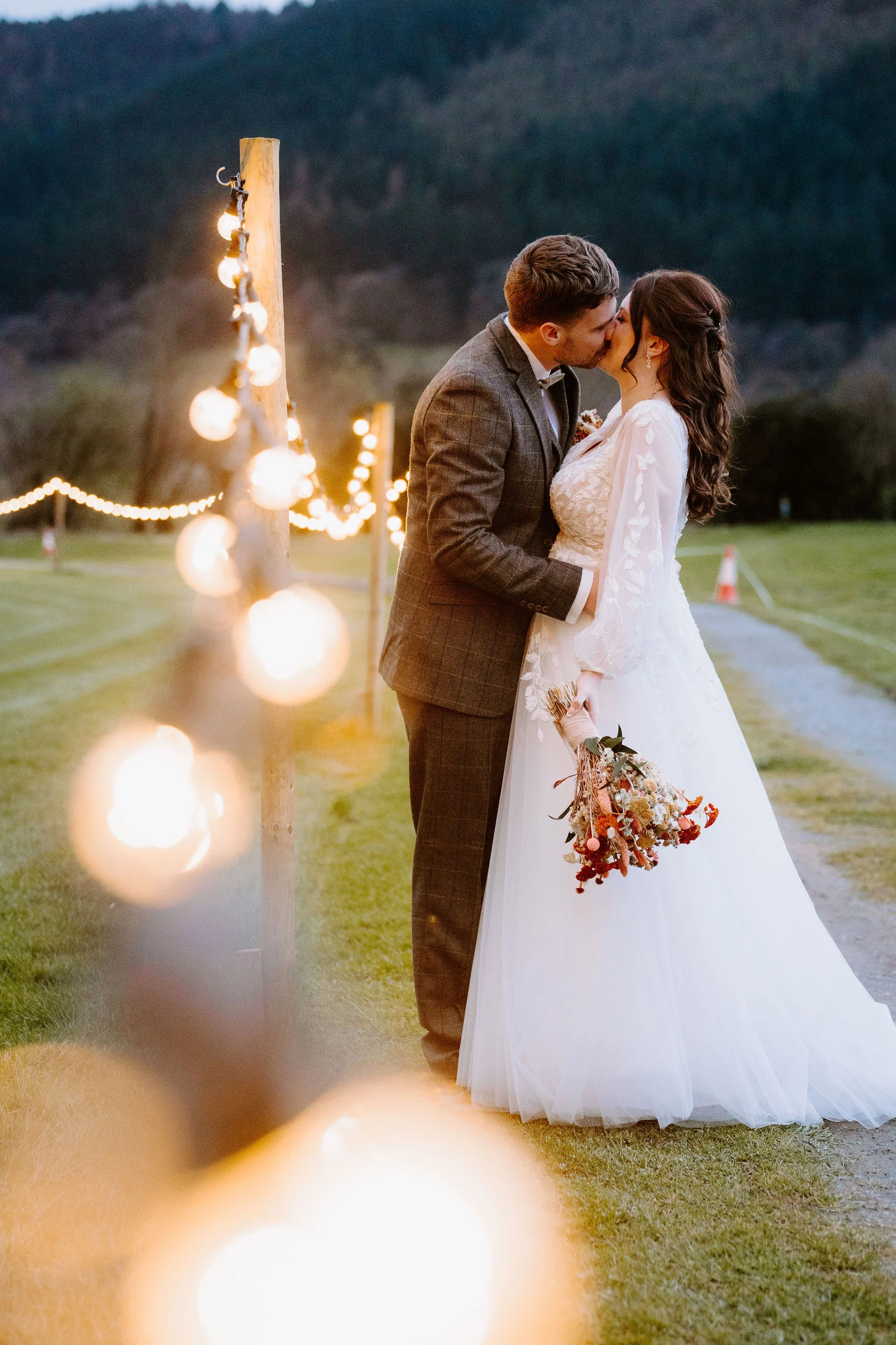 A bride and groom kiss on a grass track lined with festoon lights at dusk with a wooded hillside behind them at Hafod Farm