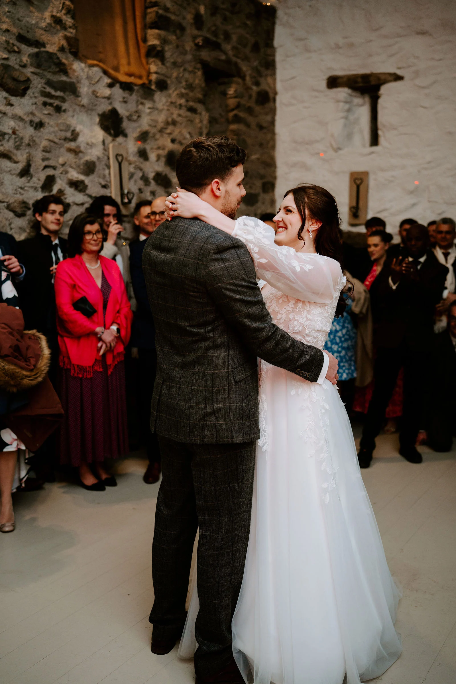 A groom in a tweed jacket holds his bride close as they dance in a whitewashed stone room with guests watching at Hafod Farm