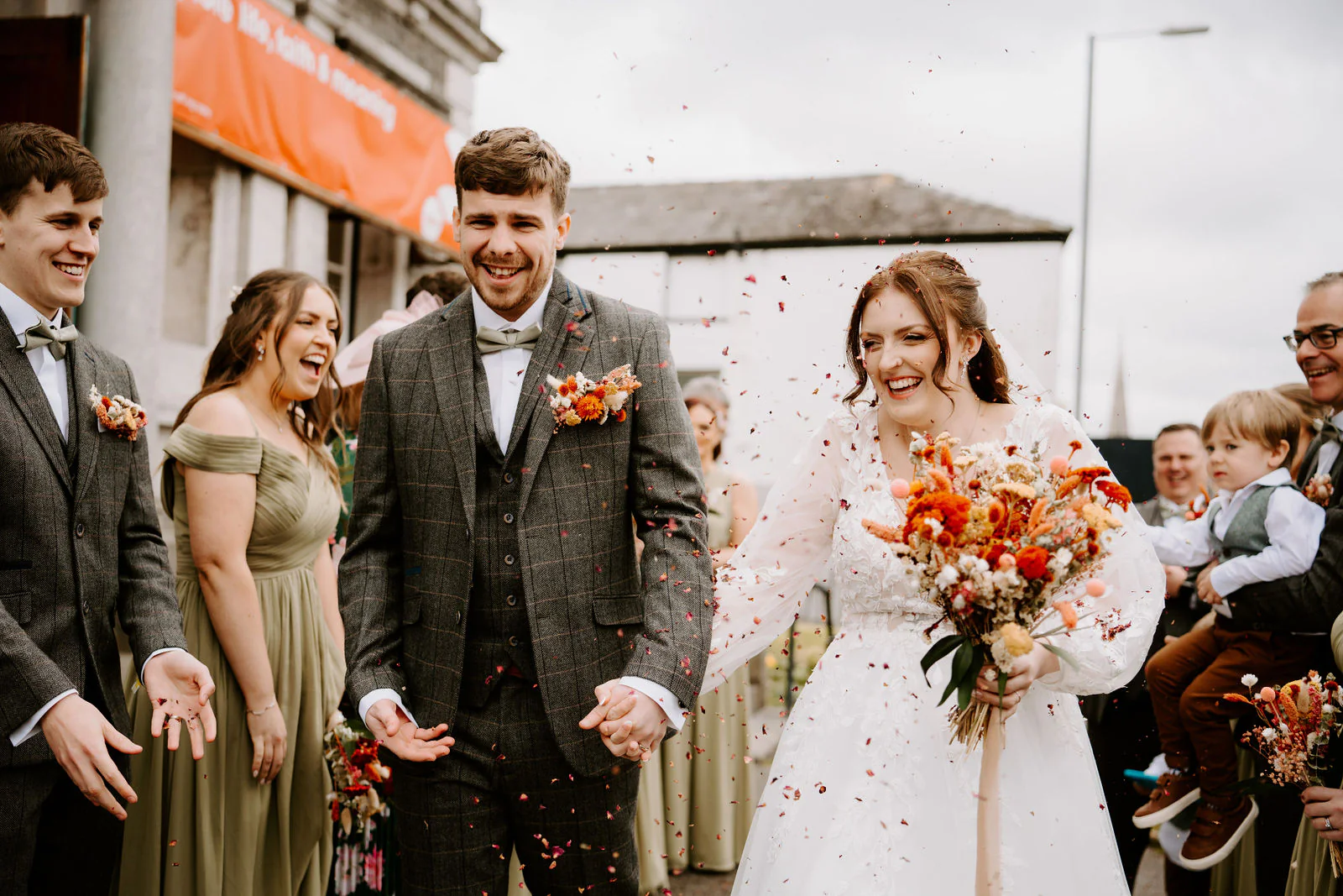 Rebecca in a long-sleeved gown and Sam in a tweed waistcoat laugh as petals fly around them in front of their bridesmaids and a small child