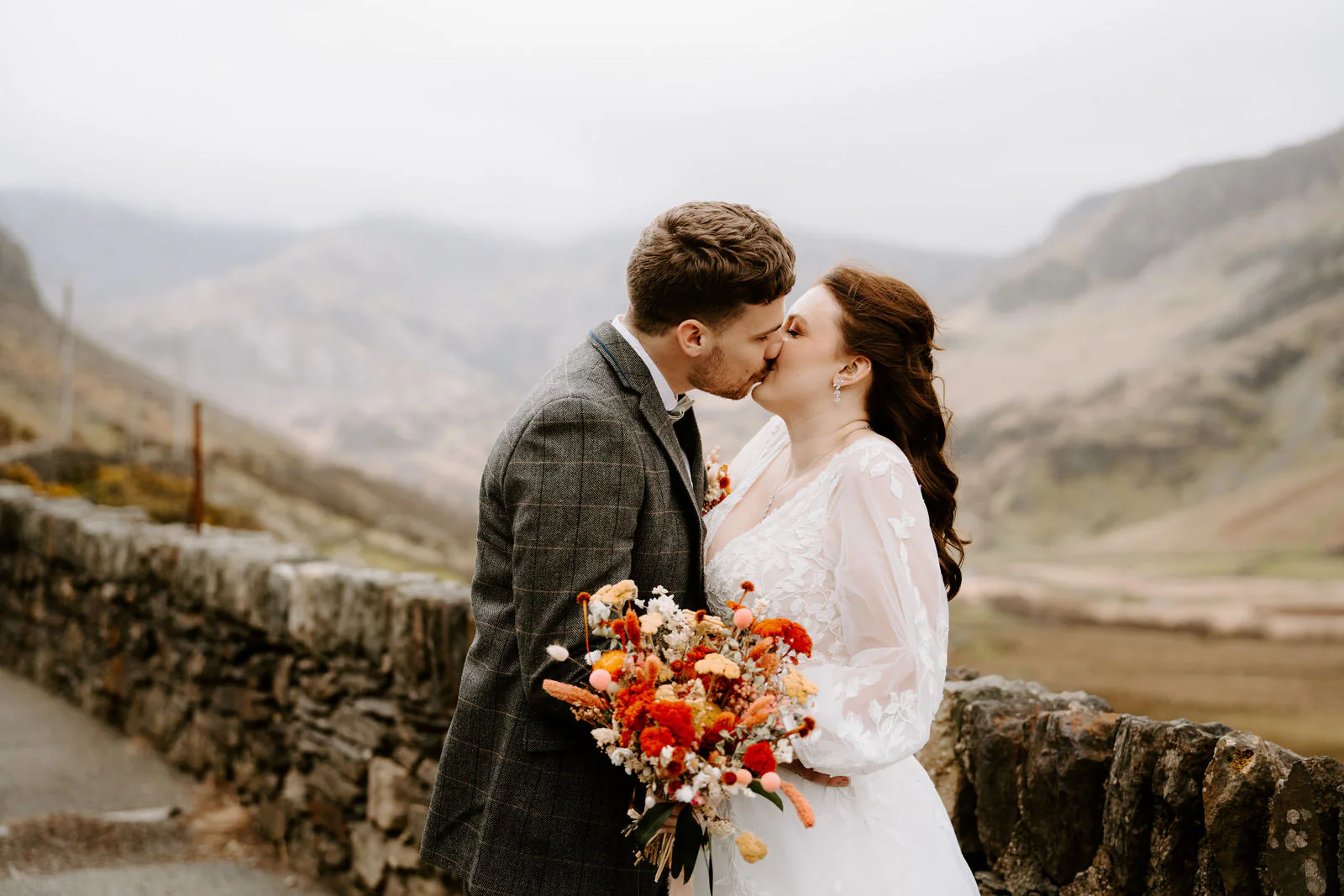 Rebecca and Sam kiss beside a dry-stone wall with a steep mountain valley rising behind them, Rebecca holding a bouquet of orange flowers