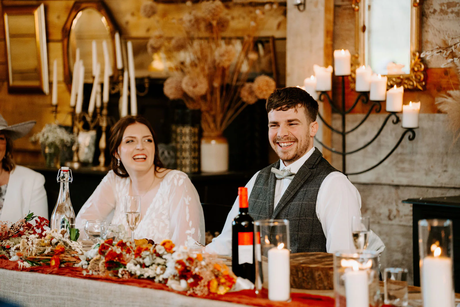 Rebecca and Sam smile at the top table in front of a mirror, dried flowers, candles and a red table runner