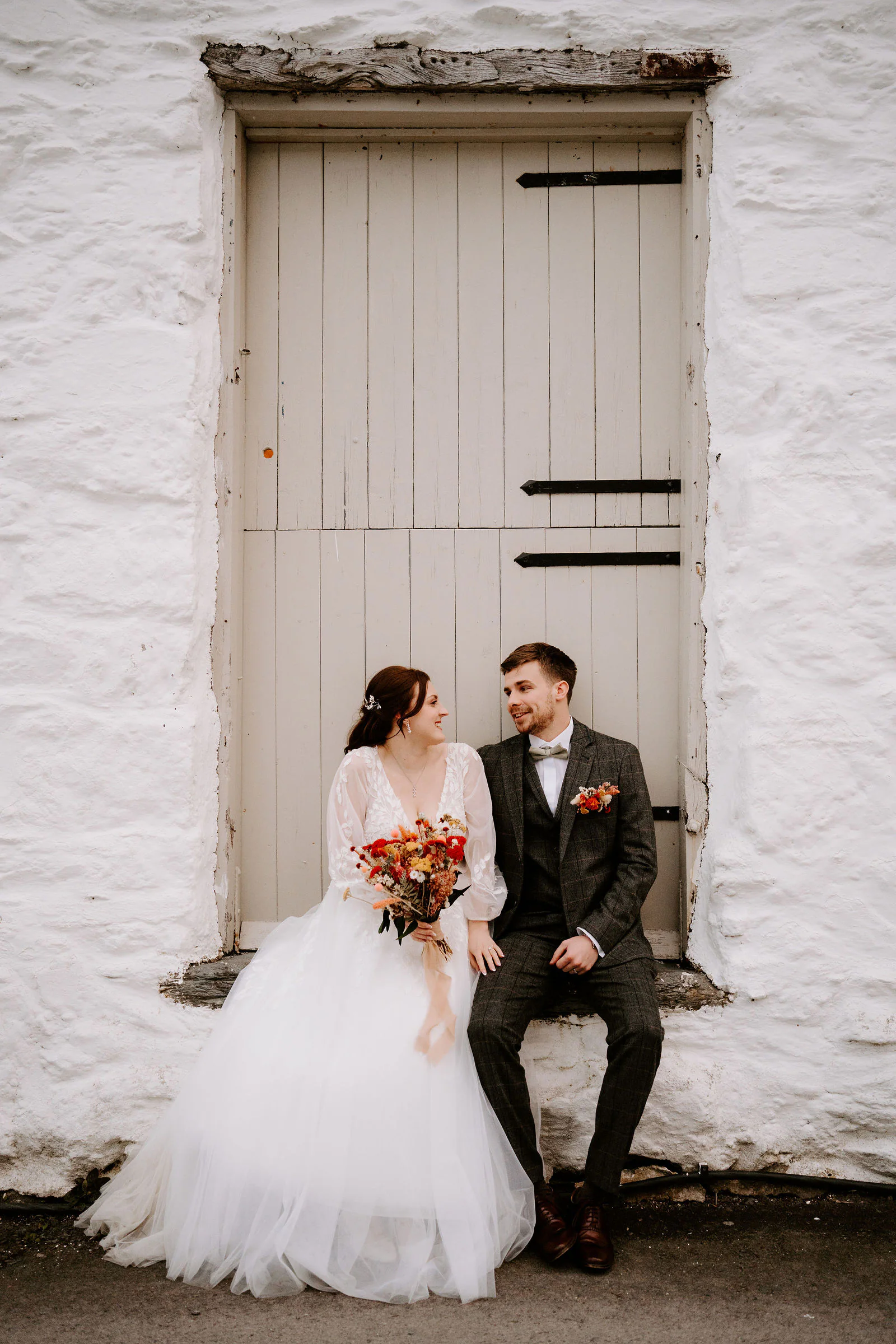 Rebecca and Sam sit together on the step of a white-washed stone building in front of a grey wooden door, Rebecca holding an orange bouquet
