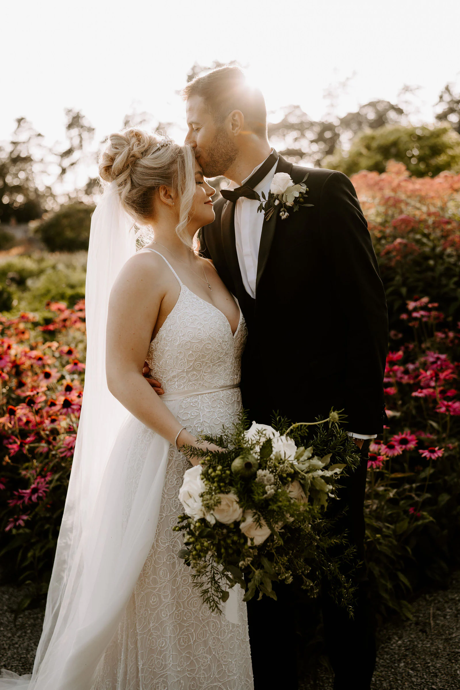 A groom kisses his bride's forehead in front of pink flowers