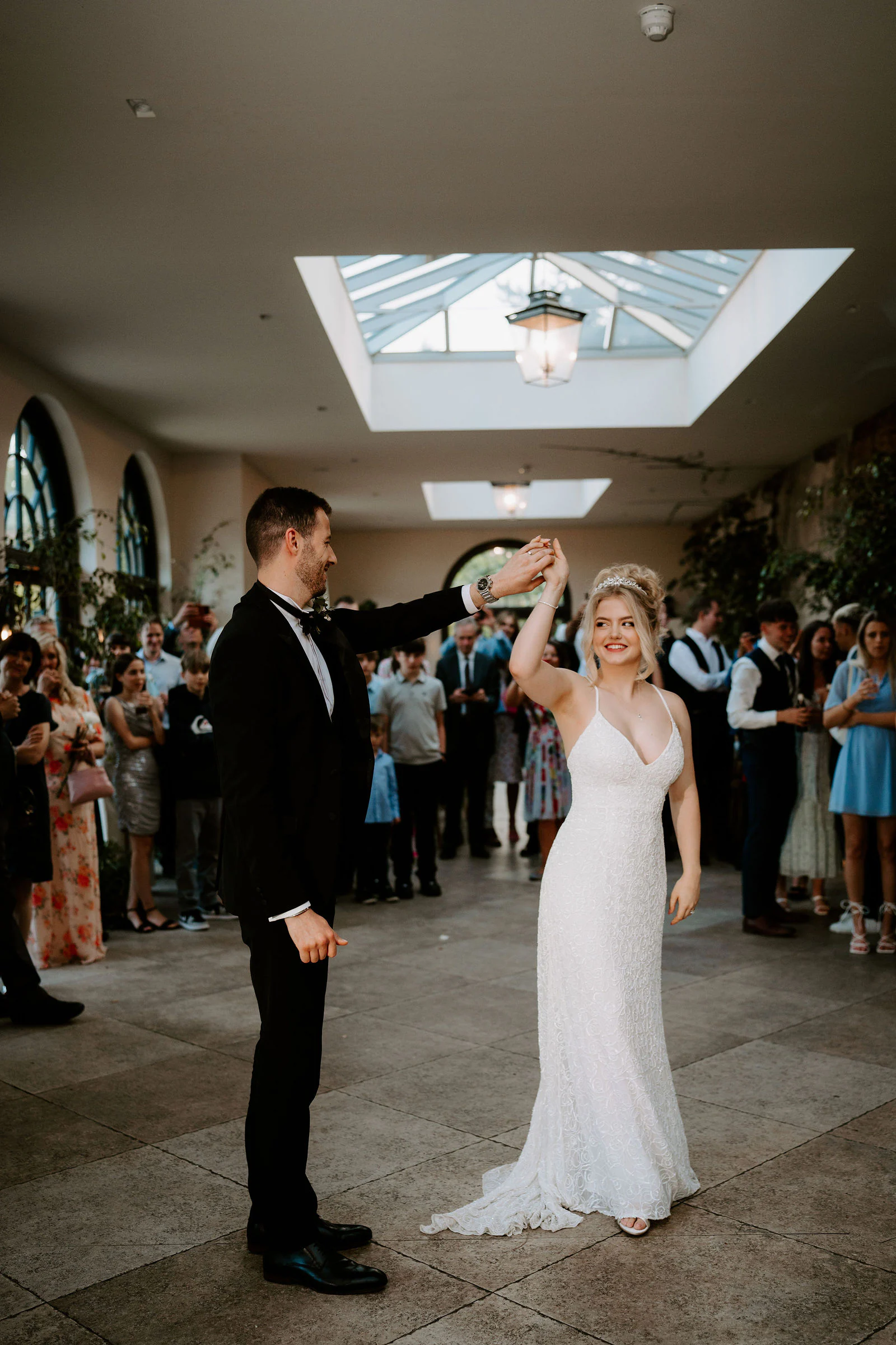 A groom twirls his bride in a stone hall while guests watch and a skylight shines above